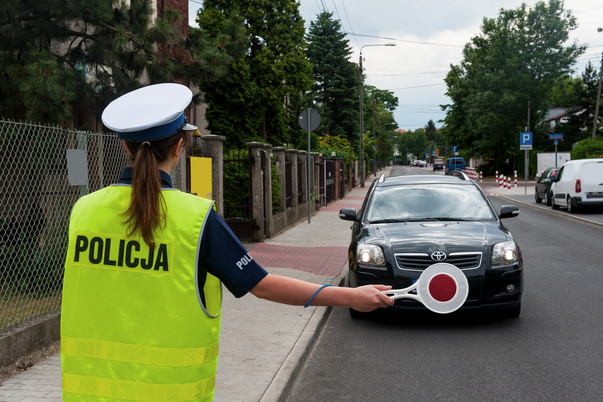 Zdjęcie przedstawia policjantkę zatrzymującą samochód do kontroli. Policjantka ustawiona jest tyłem do obserwatora zdjęcia, w wyciągniętej prawej ręce ma czerwony lizak. Zatrzymywany samochód to elegancka toyota w ciemnych barwach. Miejsce akcji to niezbyt ruchliwa droga w dzielnicy willowej.