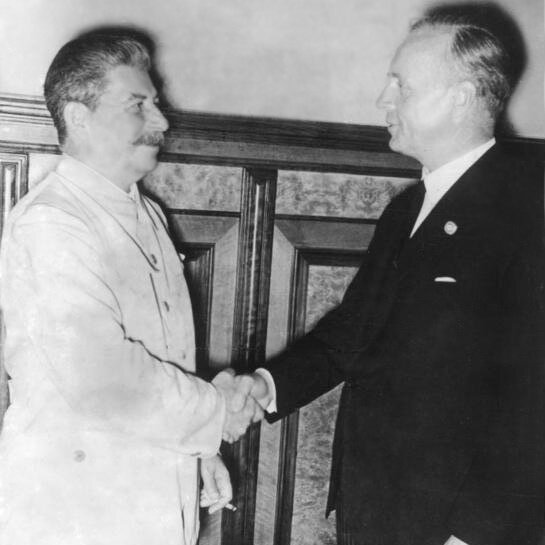 Ilustracja przedstawia dwóch mężczyzn stojących naprzeciwko siebie i ściskających sobie dłonie. Mężczyzna po lewo ubrany jest w jasną marynarkę, ma szpakowate włosy zaczesane do tyłu i wąsy. W lewej dłoni trzyma papierosa. To Józef Stalin. Mężczyzna po prawo ubrany jest w ciemny garnitur i krawat oraz jasną koszule. To Joachim von Ribbentrop.