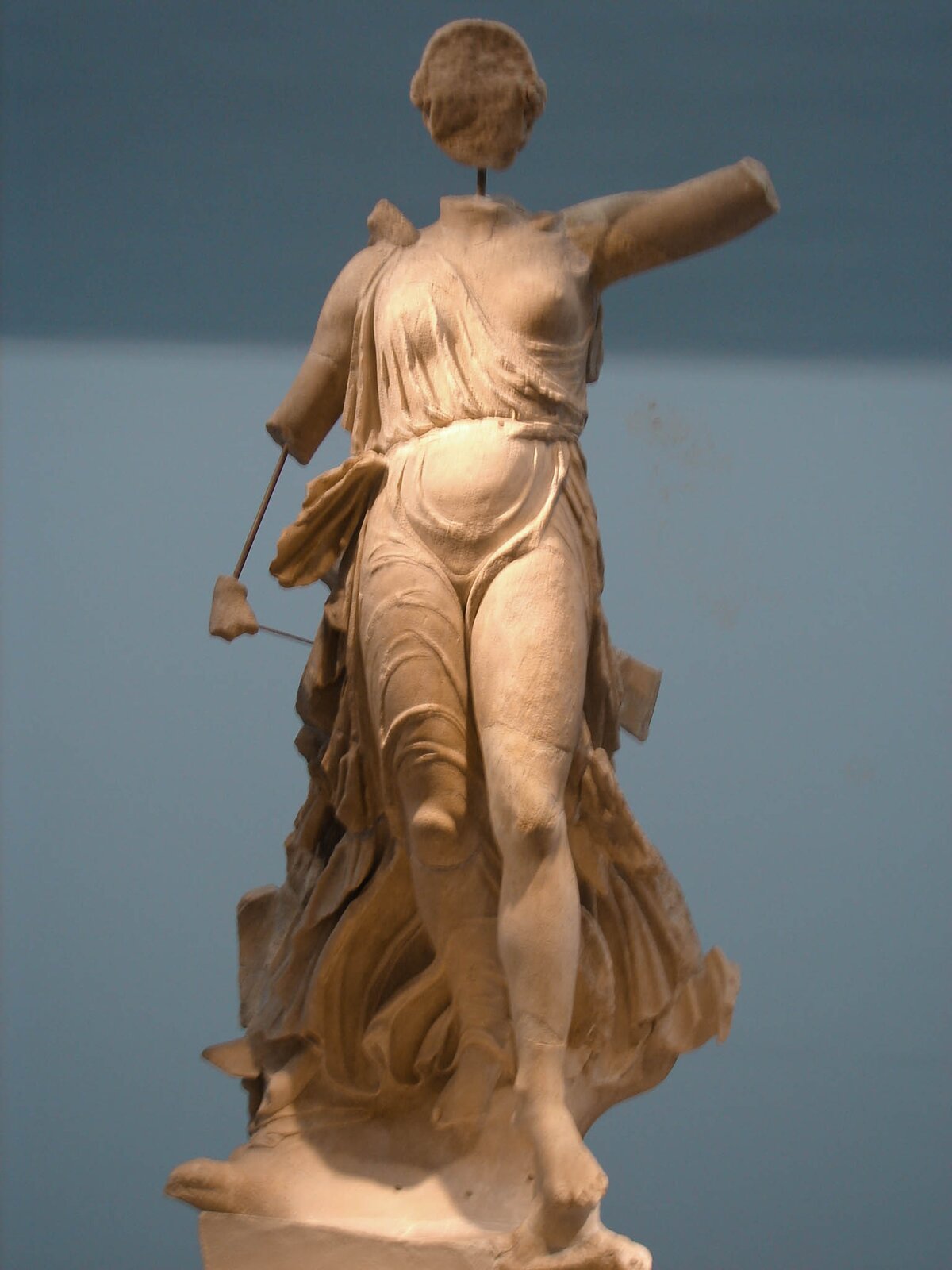 Zdjęcie przedstawia zniszczoną rzeźbę. To postać szczupłej kobiety w udrapowanej, rozwianej szacie. Kobieta jest nachylona do przodu, jakby leciała. Lewą rękę (utrąconą poniżej ramienia) ma skierowaną do przodu. Prawa ręka jest także utrącona nad łokciem. Brakuje także dużej części głowy i całej twarzy.