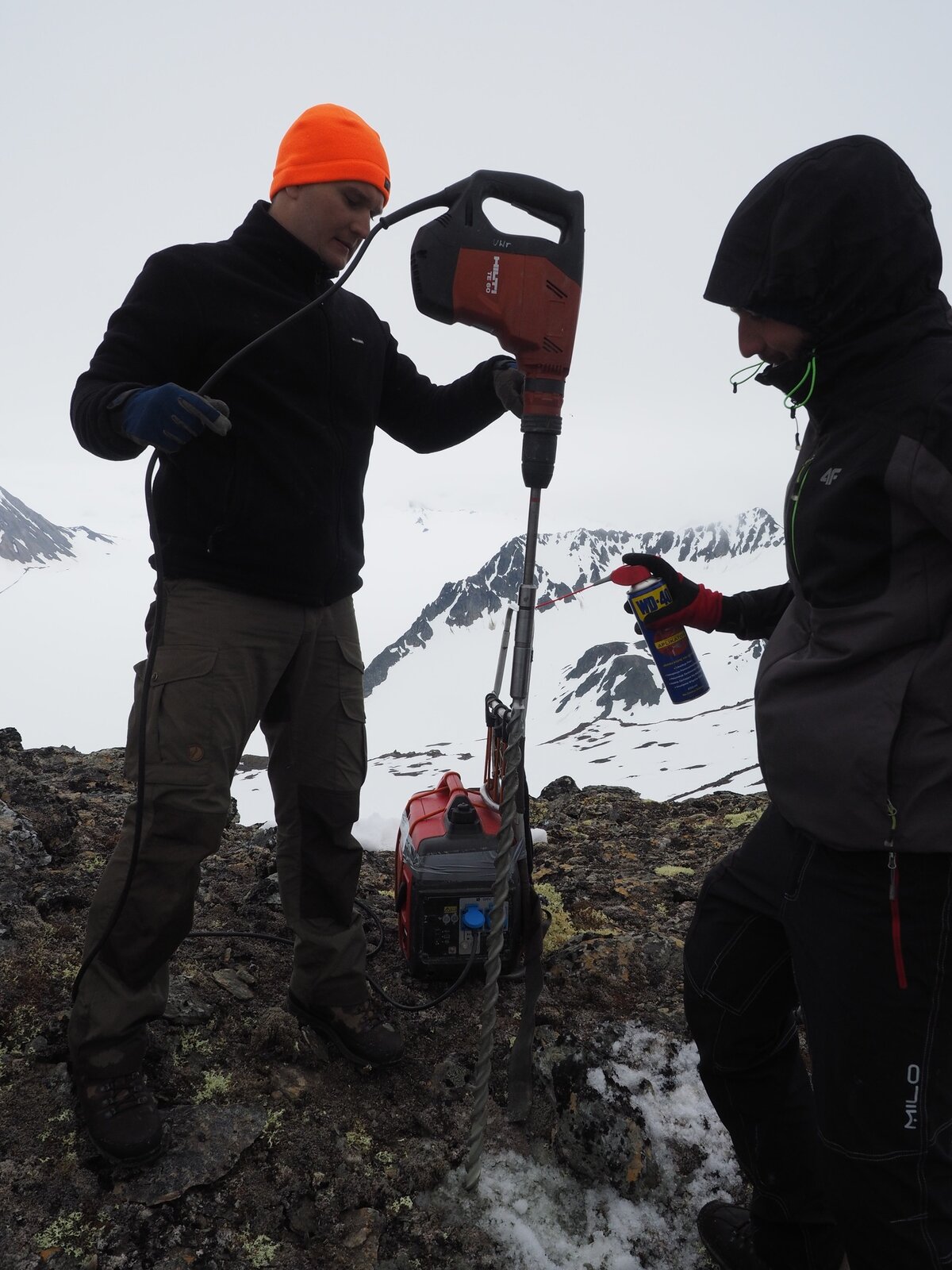 Na zdjęciu dwóch naukowców w trakcie prowadzonych badań. Prowadzą odwiert za pomocą bardzo długiego wiertła. Ziemia pod nimi pokryta gdzieniegdzie porostami, w większości pokryta śniegiem, w tle góry.