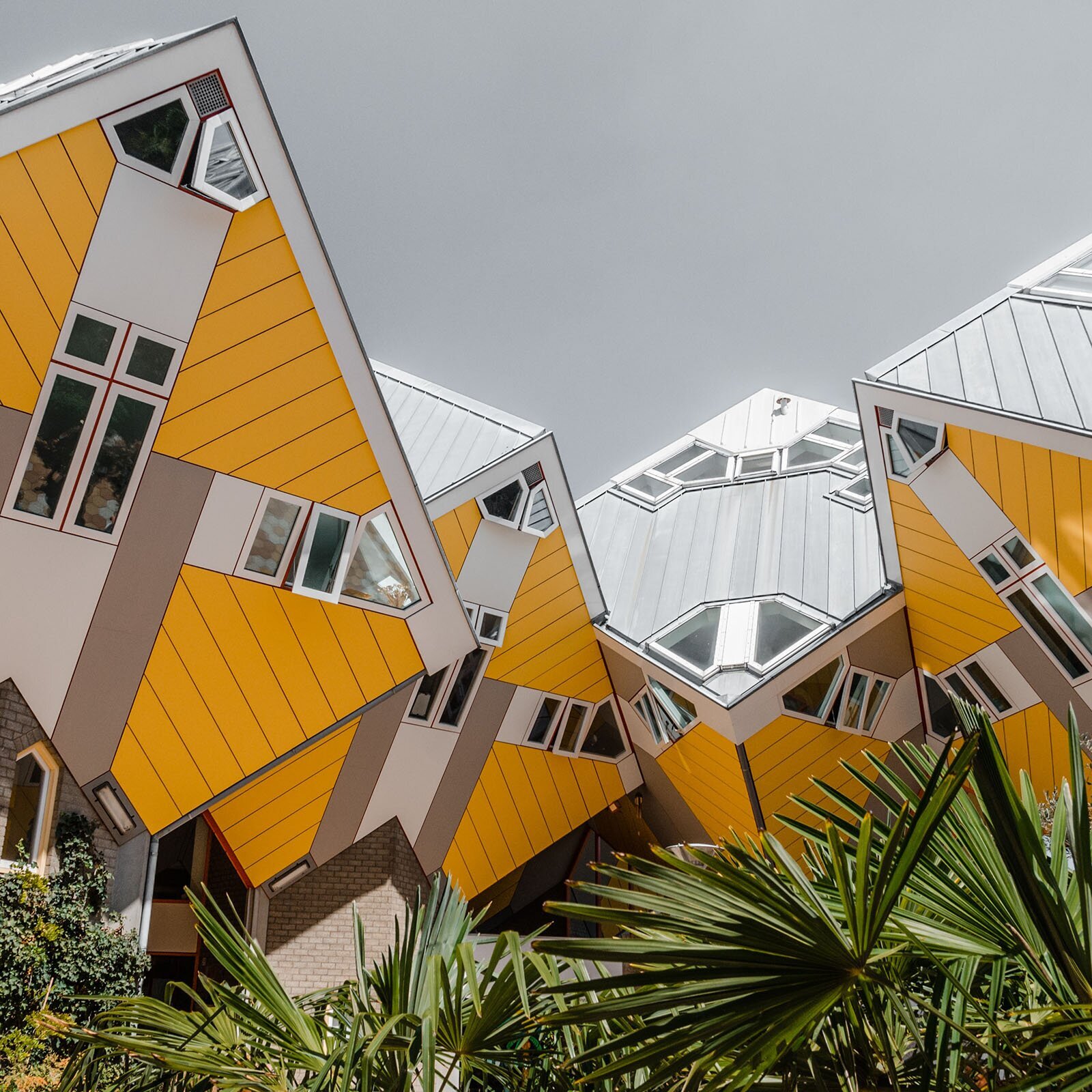 Zdjęcie przedstawia domy wykonane w stylu architektury futurystycznej. Przypominają one masywne sześciany wbite rogami w ziemię i połączone ze sobą rogami bocznymi. Ich ściany są żółto białe, a dachy szare. Posiadają białe okna na rogach i środkach każdej ściany sześcianu. Okna te otwierają się w przypadkowe strony. Przed konstrukcją rosną egzotyczne krzaki. 