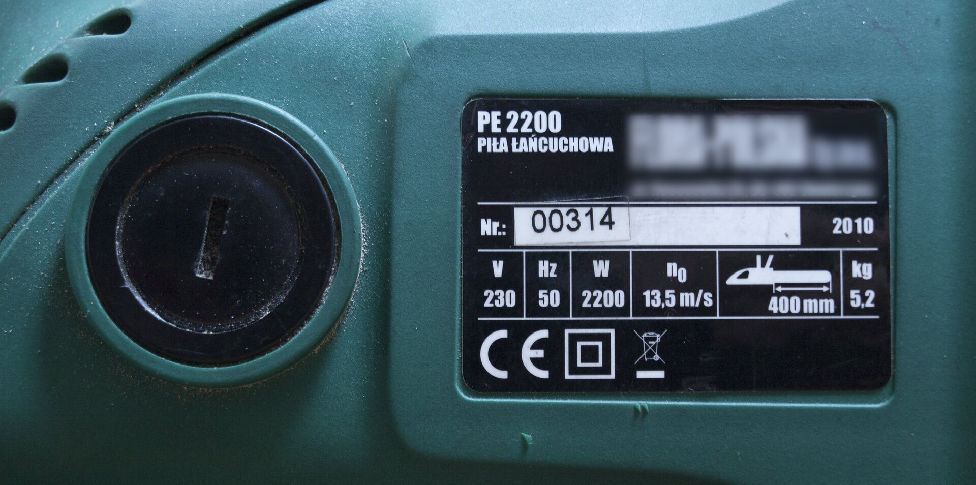 Zdjęcie przedstawia tabliczkę znamionową na obudowie elektrycznej piły łańcuchowej. Obudowa urządzenia zielona, po lewej stronie zdjęcia czarna zakrętka szczotki silnika używana podczas jej wymiany. Po prawej stronie czarna tabliczka z symbolem towaru, typem (piła łańcuchowa), numerem seryjnym i rokiem produkcji. Poniżej podstawowe parametry urządzenia: napięcie 230 woltów, 50 herców, moc 2200 watów, prędkość przesuwu łańcucha 13,5 metra na sekundę, długość prowadnicy 400 milimetrów, masa 5,2 kilograma.