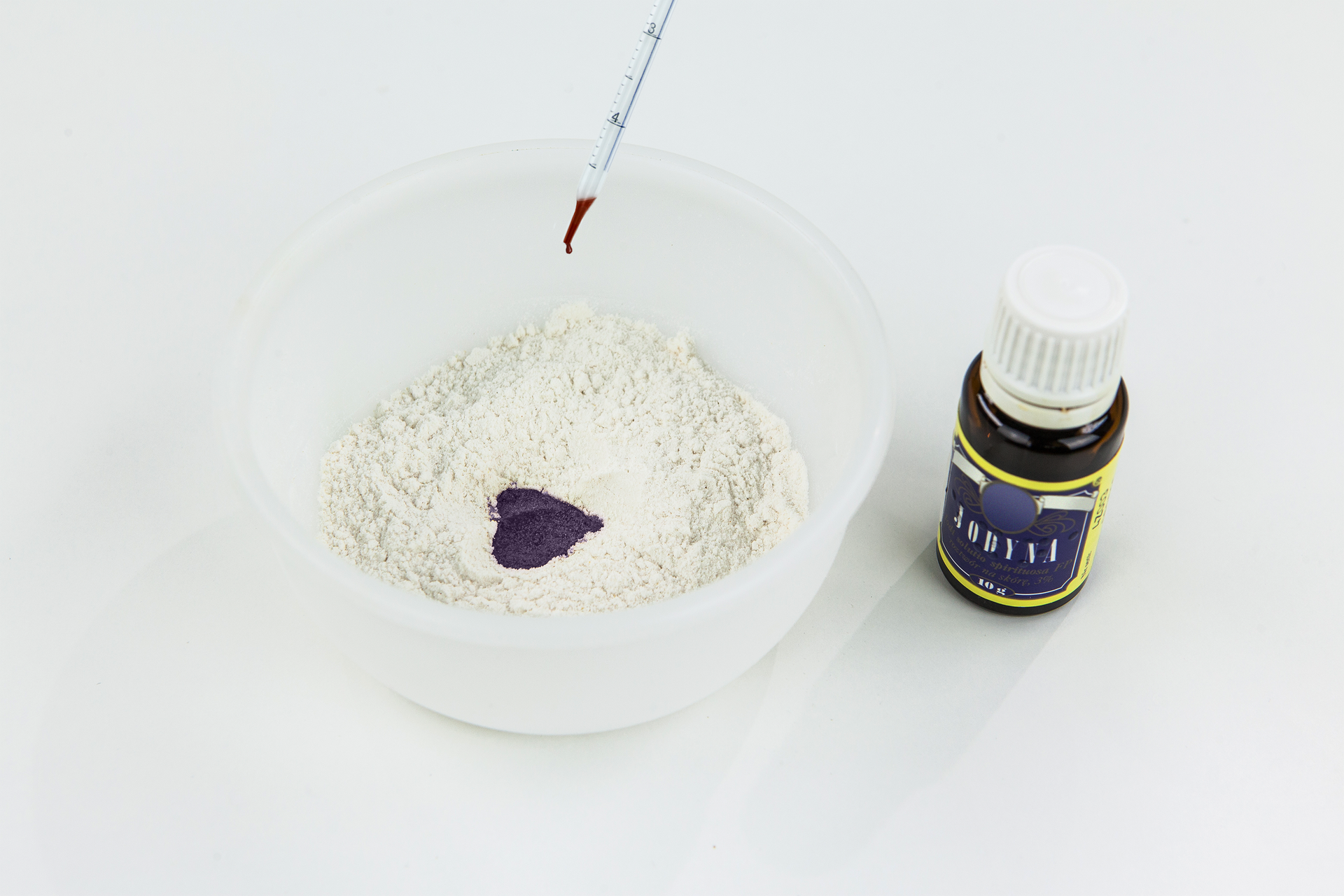 Fotografia przedstawia na białym tle pojemnik z białą mąką. Obok stoi brązowa buteleczka z białym korkiem i granatową etykietą z napisem: jodyna. Nad mąka szklana pipetka miarowa z brązową cieczą u dołu. Jej kropla zaraz spadnie. Pod pipetką na mące granatowa, sercowata plama. W ten sposób wykrywa się skrobię.