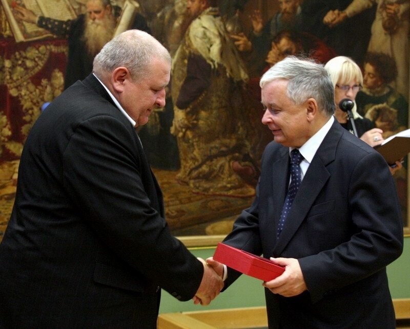 W pomieszczeniu dwaj mężczyźni podający sobie prawe dłonie. Po lewej stronie stoi Marek Franczak, syn żołnierza wyklętego. Po prawej stronie stoi prezydent Lech Kaczyński. W lewej dłoni trzyma pudełko, w którym znajduje się odznaczenie. 