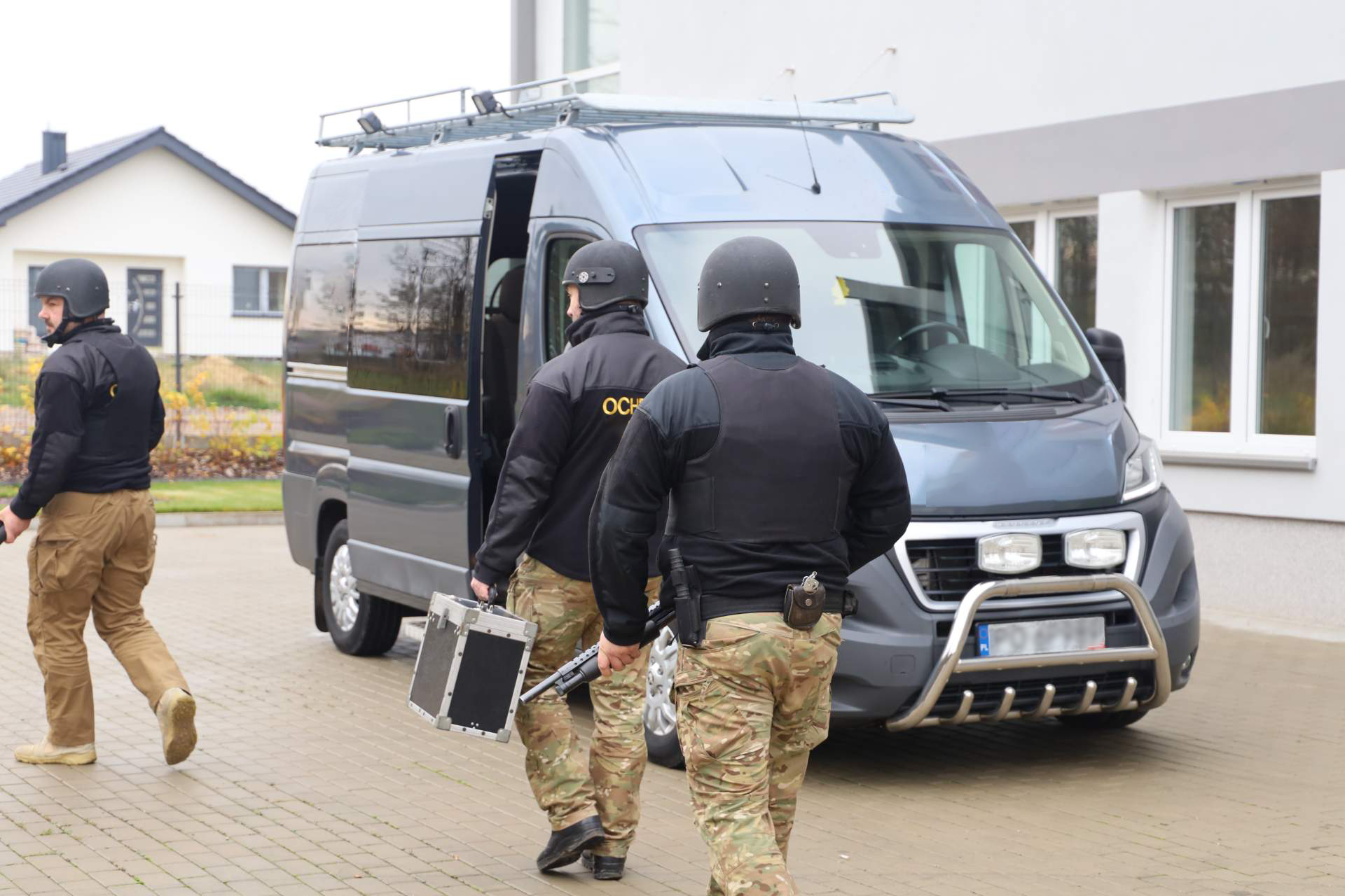 Krok trzeci. Przygotowanie obiektu oraz przylegającego terenu. Zdjęcie czwarte, przedstawia trzech ochroniarzy w spodniach z kamuflażem, czarnym polarze, oraz kamizelkach kuloodpornych. Mężczyźni transportują walizkę, ochraniając ją przy użyciu strzelb. Przed mężczyznami znajduje się w furgonetka z otwartymi drzwiami.