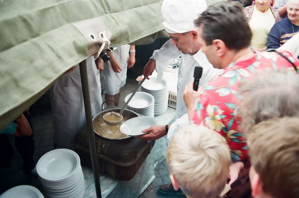 Zdjęcie przedstawia mężczyznę ubranego w biały fartuch i czapkę kucharską, który chochlą nalewa zupę z dużego gara na talerz. obok niego mężczyzna z mikrofonem, wokół czekający ludzie. Obok kotła z zupą stoją talerze.
