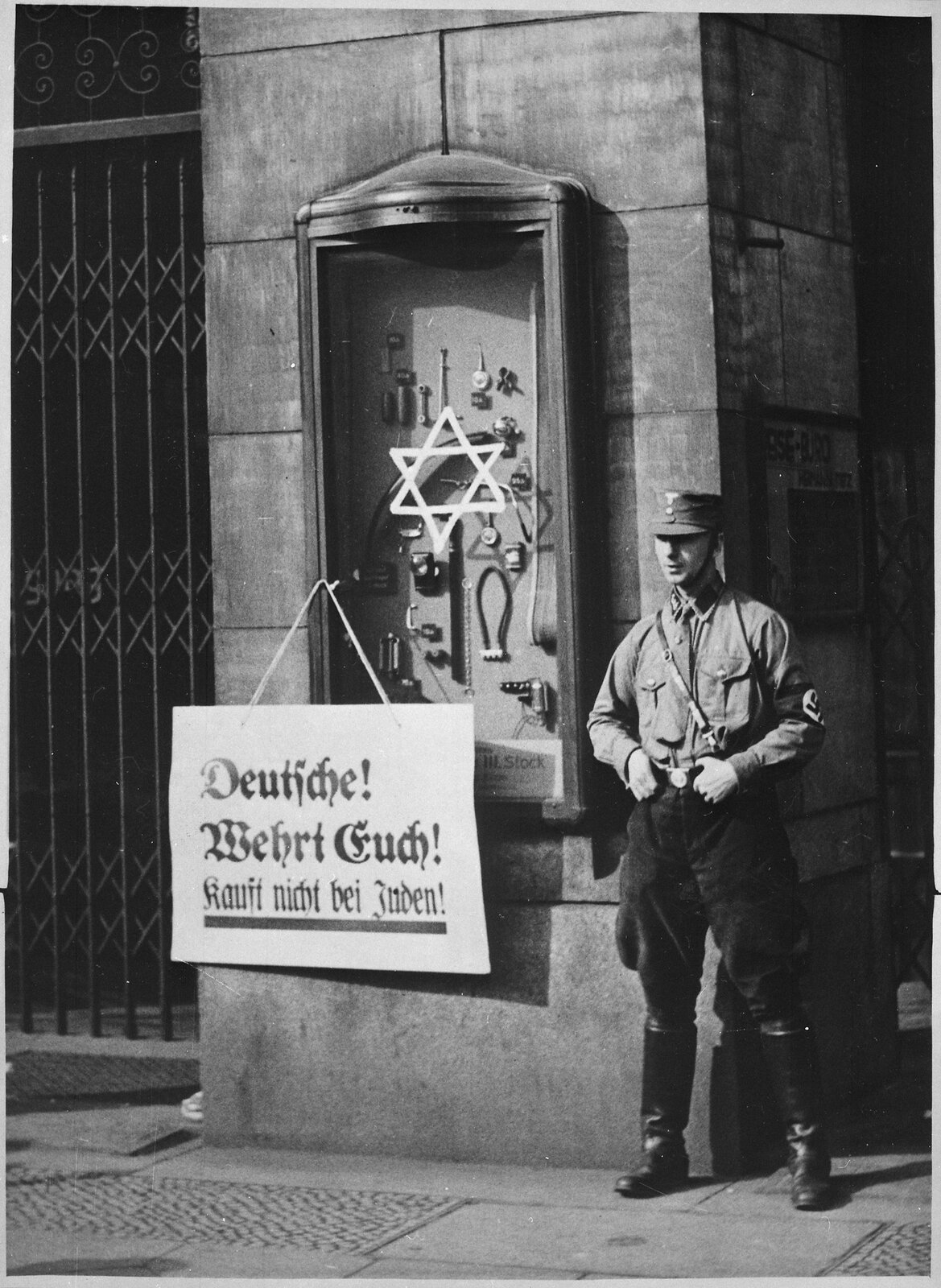 Zdjęcie przedstawia gablotę z reklamą warsztatu rzemieślniczego prowadzonego przez Żydów. Na gablocie namalowana jest gwiazda Dawida. Obok gabloty stoi mężczyzna w mundurze NSDAP. Na gablocie wisi informacja w języku niemieckim.