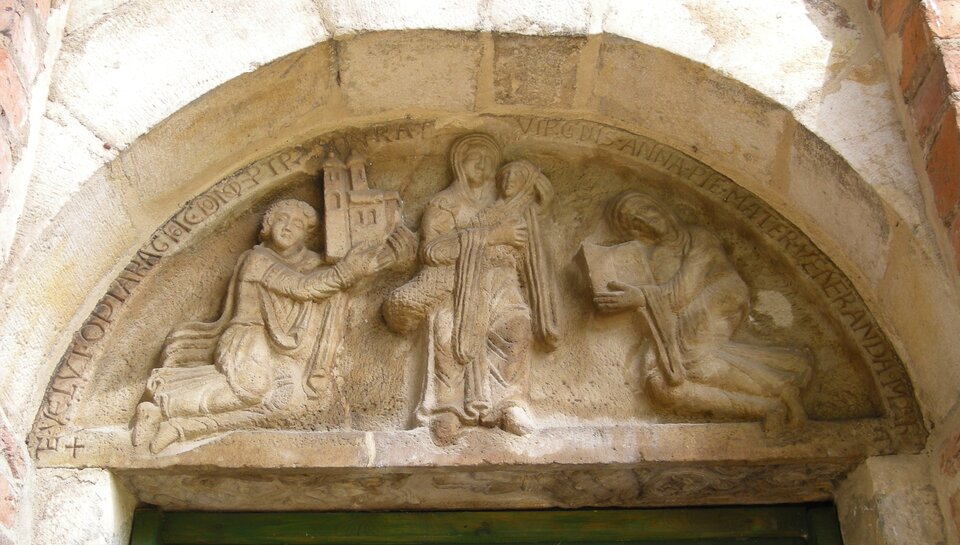 Ilustracja o kształcie poziomego prostokąta przedstawia fotografię tympanonu fundacyjnego z kościoła św. Trójcy w Strzelnie. Zdjęcie ukazuje św. Annę trzymającą małą Marię. Z lewej strony klęczy mężczyzna, w rękach trzyma model świątyni. Z prawej strony klęczy kobieta z księgą w rękach. Pozostałą część zdjęcia wypełnia fragment ściany. 