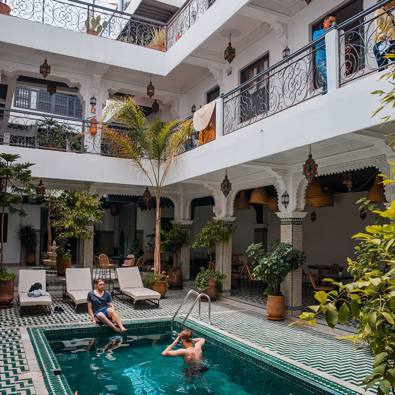 Zdjęcie przedstawia przytulny hol piętrowego budynku. Po środku holu umieszczono mały basen, wyłożony kafelkami w kolorach bieli i zieleni. W basenie znajduje się dwóch młodych mężczyzn, a dookoła rozstawiono tropikalne rośliny w glinianych donicach i białe leżaki. Na pierwszym i drugim piętrze dostrzegamy prostokątne przejścia otoczone barierkami. 