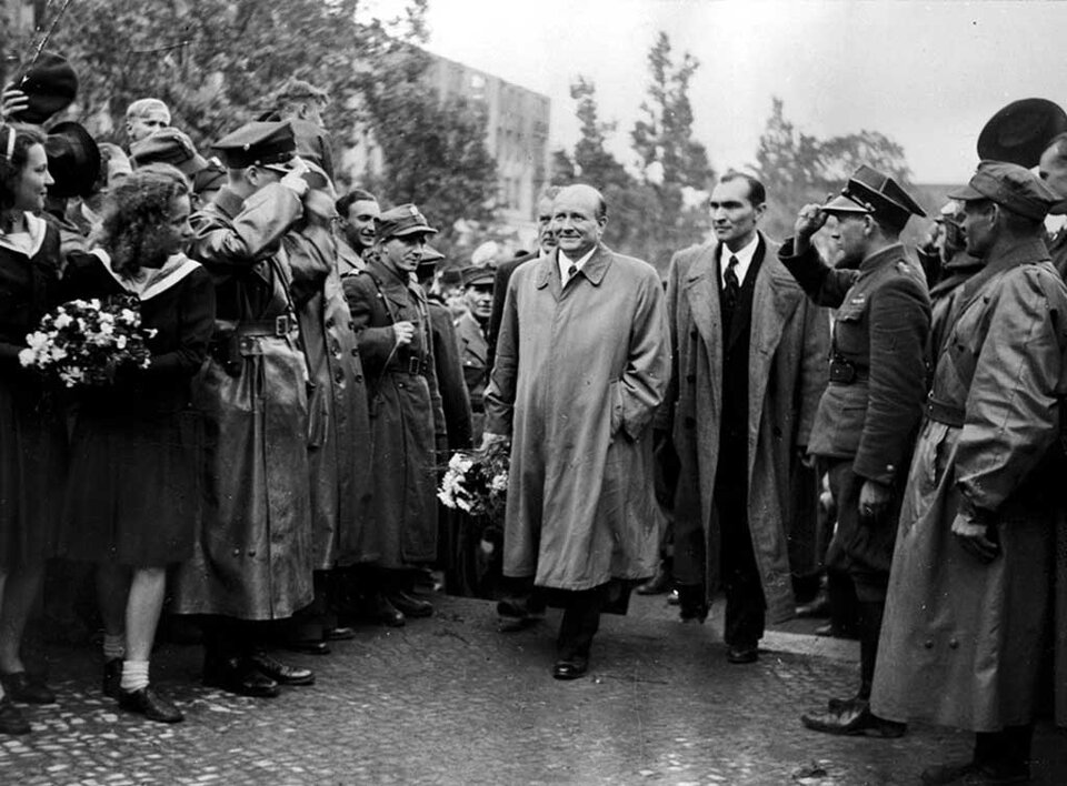 Zdjęcie przedstawia grupę ludzi: żołnierzy,  cywilów i młodych harcerek, którzy radośnie witają przechodzącego pomiędzy nimi mężczyznę. Idzie on w towarzystwie dwóch innych, eskortujących go osób, które są ubrane podobnie jak on. Mężczyzna idący w środku ma w ręku kwiaty. Jest średniego wzrostu, łysiejący, ma okrągłą  i pogodną twarz. Ubrany jest w koszulę, krawat, spodnie, eleganckie buty i długi zapięty płaszcz. Pośród zebranych w szpalerze żołnierzy dwóch salutuje. Jeden z nich ubrany jest w mundur, a drugi w długi wojskowy płaszcz, obaj mają czapki rogatywki. Niektórzy  z zebranych podnoszą do góry, w geście radości, nakrycia głowy, a harcerki trzymają w ręku kwiaty. W oddali widać drzewa i wysoki, prostokątny budynek.    