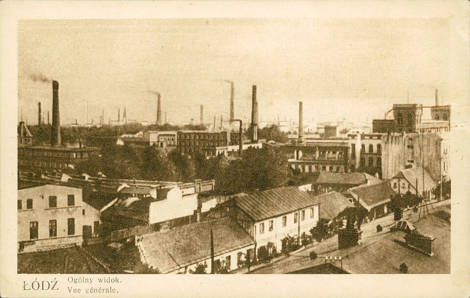 Zdjęcie przedstawia panoramę miasta. Wśród budynków znajdują się wysokie kominy, z których wydobywa się dym.