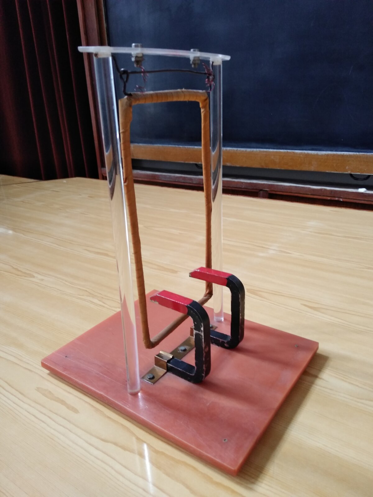 Rys. a. Na stole ustawiono obok siebie 2 magnesy o kształcie podkowy. Magnesy ustawione są równolegle do siebie,  w płaszczyźnie pionowej.  Między biegunami magnesów znajduje się dolna krawędź ramki, zawieszonej za górną krawędź, która jest osią obrotu ramki.