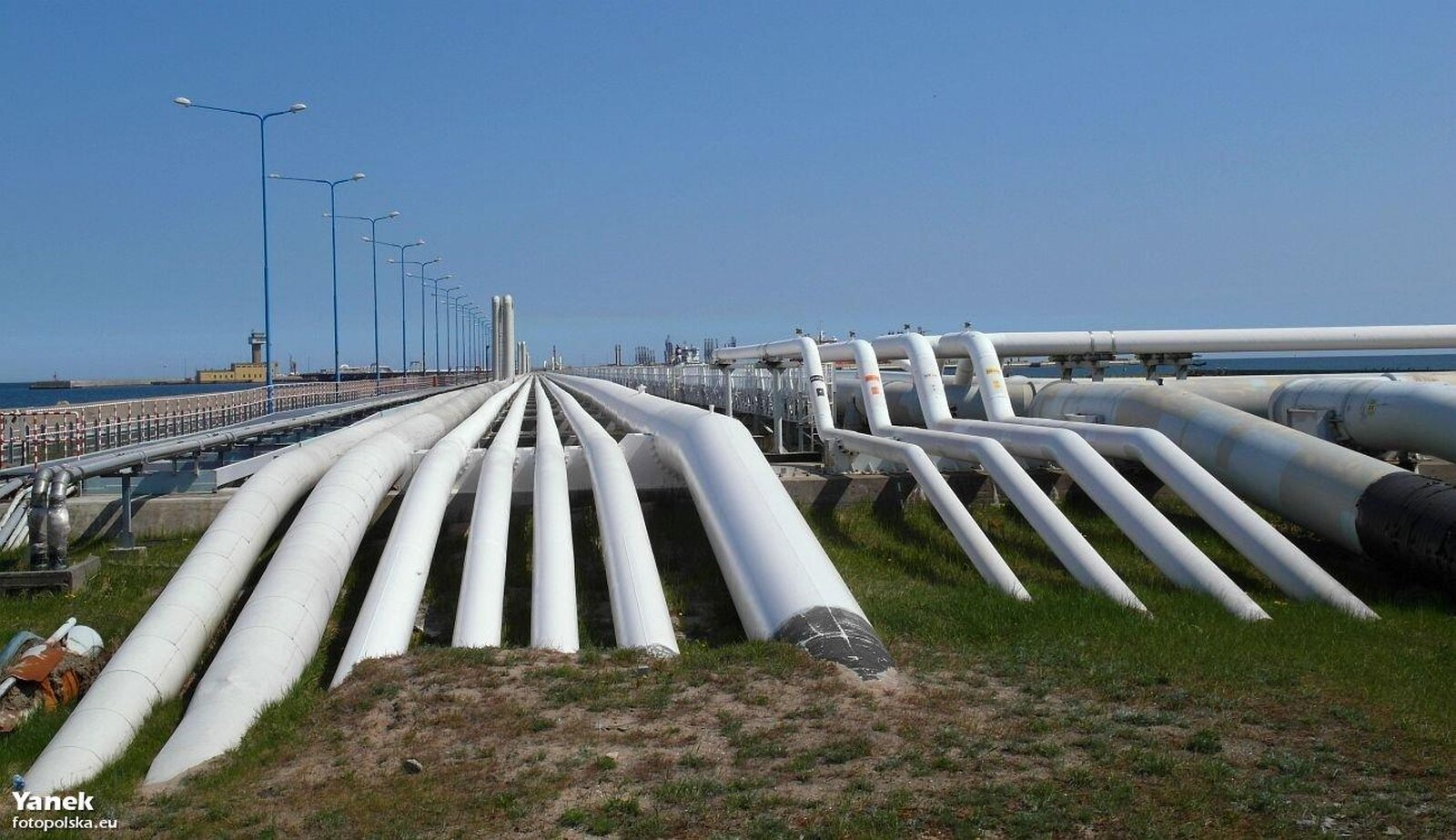 Fotografia przedstawia obszar przemysłowy. W centralnej części rury przesyłowe różnych rozmiarów. Po lewo ogrodzenie i lampy. W oddali woda. 