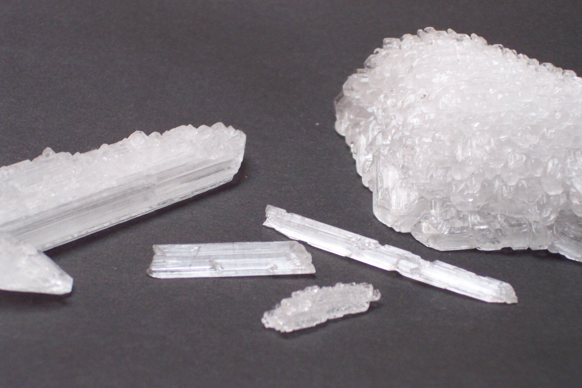 Zdjęcie przedstawiające białawe, półprzeźroczyste kryształy etanianu sodu. Mają one postać długich, grubych igieł lub pozrastanych kryształów tworzących większe struktury o nieokreślonym kształcie.