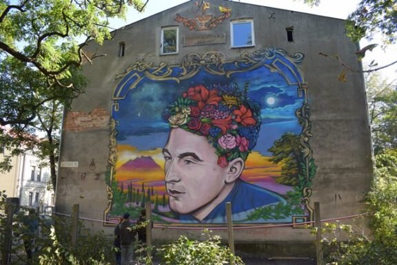 Zdjęcie przedstawia mural znajdujący się na ścianie kamienicy. Na muralu przedstawiona jest głowa młodego mężczyzny w pozycji  en trois quarts  [czytaj: trła katr]. Zamiast włosów ma różnokolorowe kwiaty. W tle znajdują się kolorowe góry i nocne niebo z księżycem.