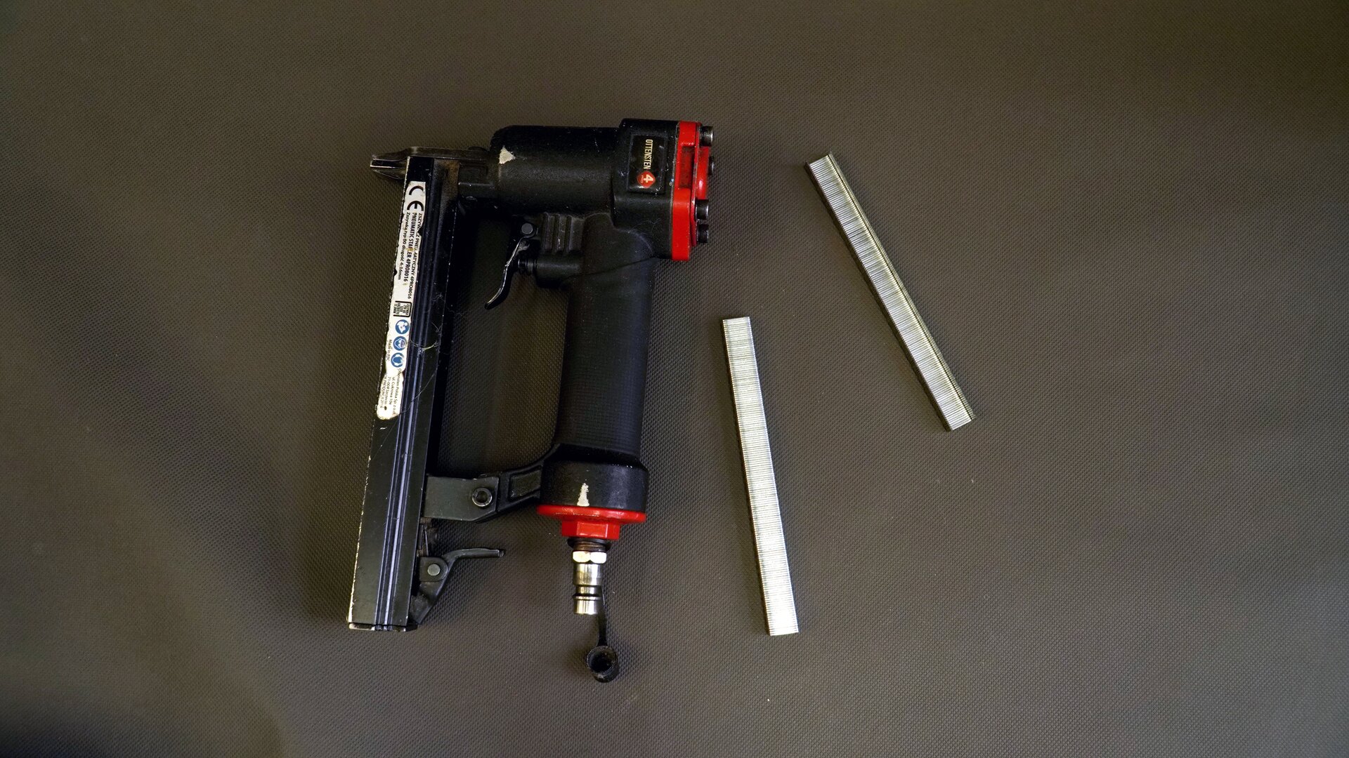 Zdjęcie przedstawia zszywacz tapicerski. Urządzenie wyposażone jest w rękojeść z uchwytem oraz podajnik zszywek. Zszywacz podłączany jest do kompresora.