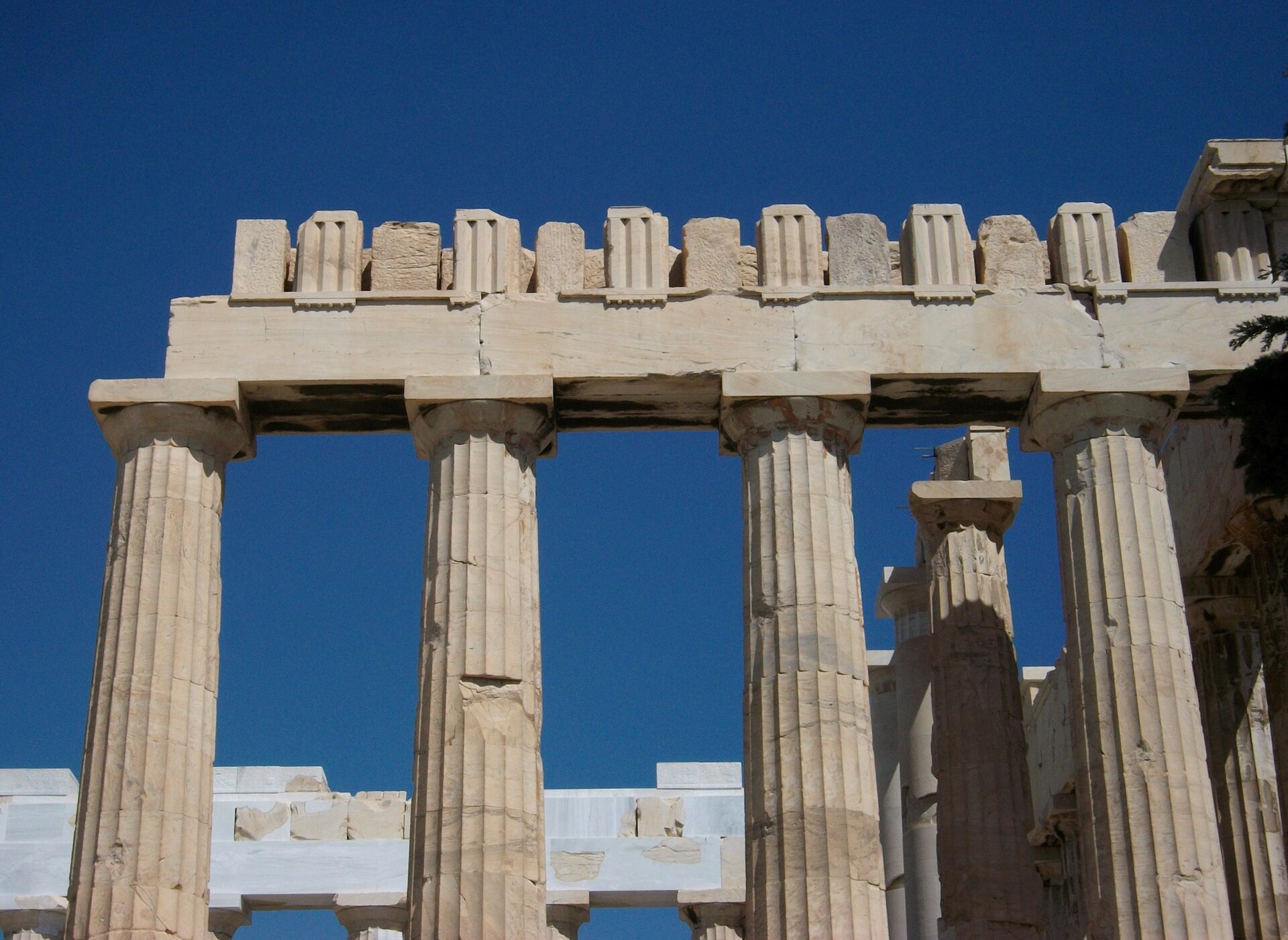 Zdjęcie przedstawia fragment kolumnady Partenonu na tle bezchmurnego nieba. Ukazana jest górna część kolumn z mocno zniszczonym belkowaniem. Kolumny są żłobkowane.