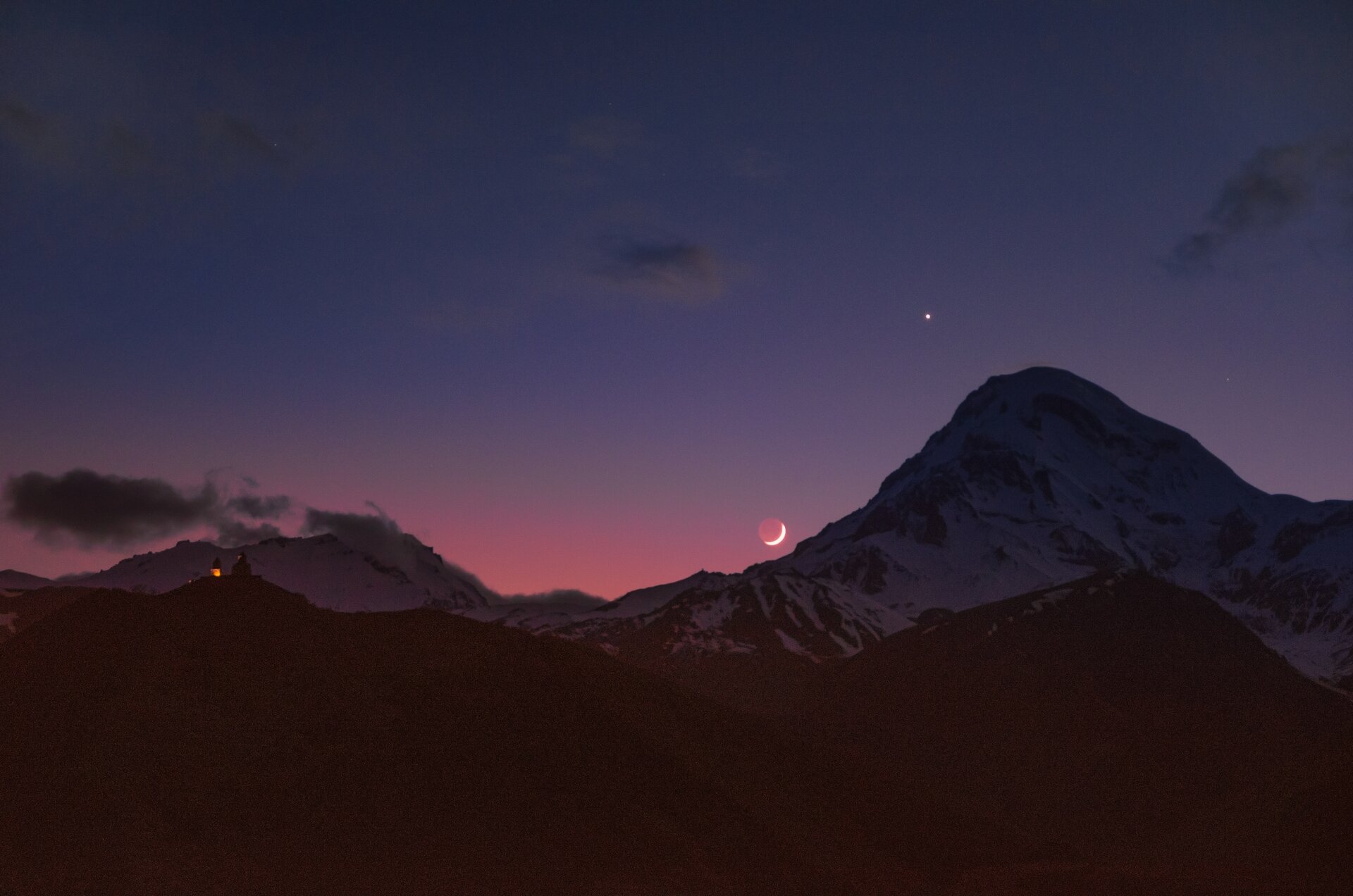 Zdjęcie przedstawia niebo widziane znad gór. Podczas zmierzchu widać na niebie księżyc i Wenus.