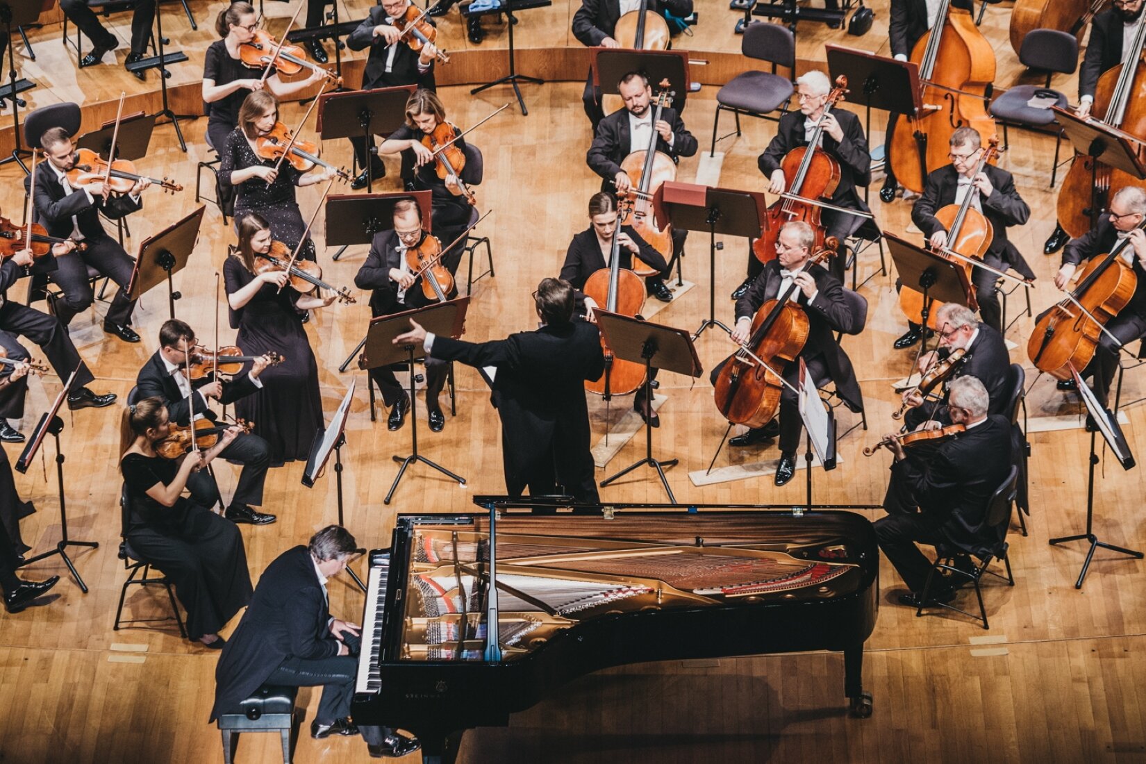 Zdjęcie przedstawia scenę sali orkiestrowej. Na pierwszym planie znajduje się mężczyzna, grający na fortepianie. Dalej ukazany jest dyrygent, stojący tyłem do widza. Jego lewa ręka jest lekko uniesiona w górę. Dalej, przedstawione są pozostałe osoby z orkiestry. Osoby te siedzą na krzesłach, grają na skrzypcach oraz wiolonczeli. Wszystkie osoby ze zdjęcia ubrane są na galowo, ich strój tworzą czarne garnitury lub sukienki. 