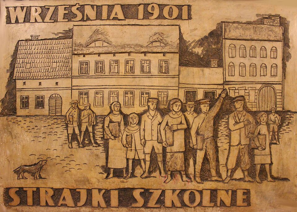 Zdjęcie przedstawia płaskorzeźbę. Prezentuje ona dzieci, które trzymają w rękach teczki. Dziewczynki mają sukienki i chusty na głowach, a chłopcy mundurki z czapkami. Jeden z chłopców unosi rękę. Za postaciami stoi rząd kamienic. Na górze jest napis: Września 1901. Na dole jest napis: Strajki szkolne.