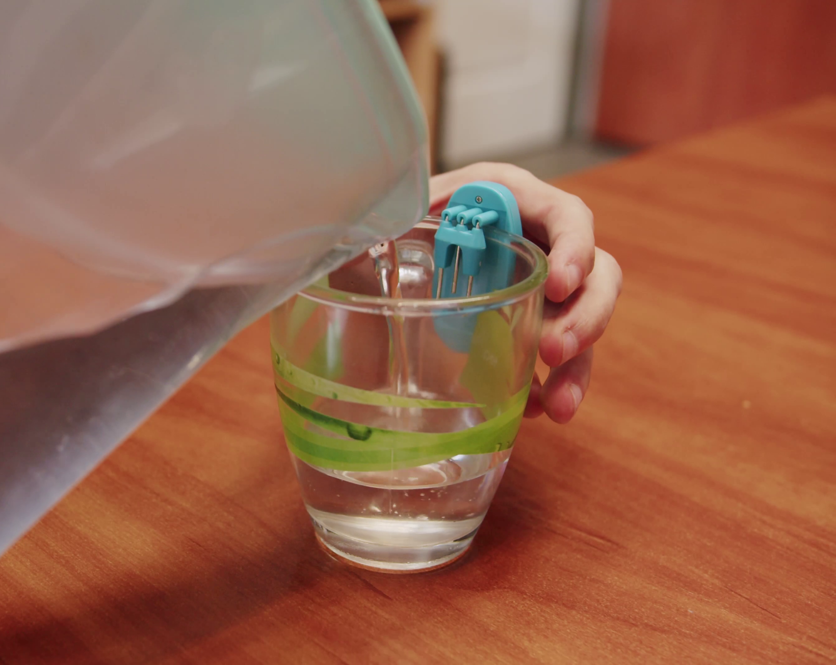 Zdjęcie przedstawia zbliżenie na szklankę trzymaną przez osobą, która nalewa do środka wodę z dzbanka. Na szklance zamontowany jest wskaźnik poziomu cieczy. To urządzenie w obudowie z tworzywa sztucznego, posiadające metalowe druciki, które aktywują dźwięk w momencie kontaktu z wodą. 