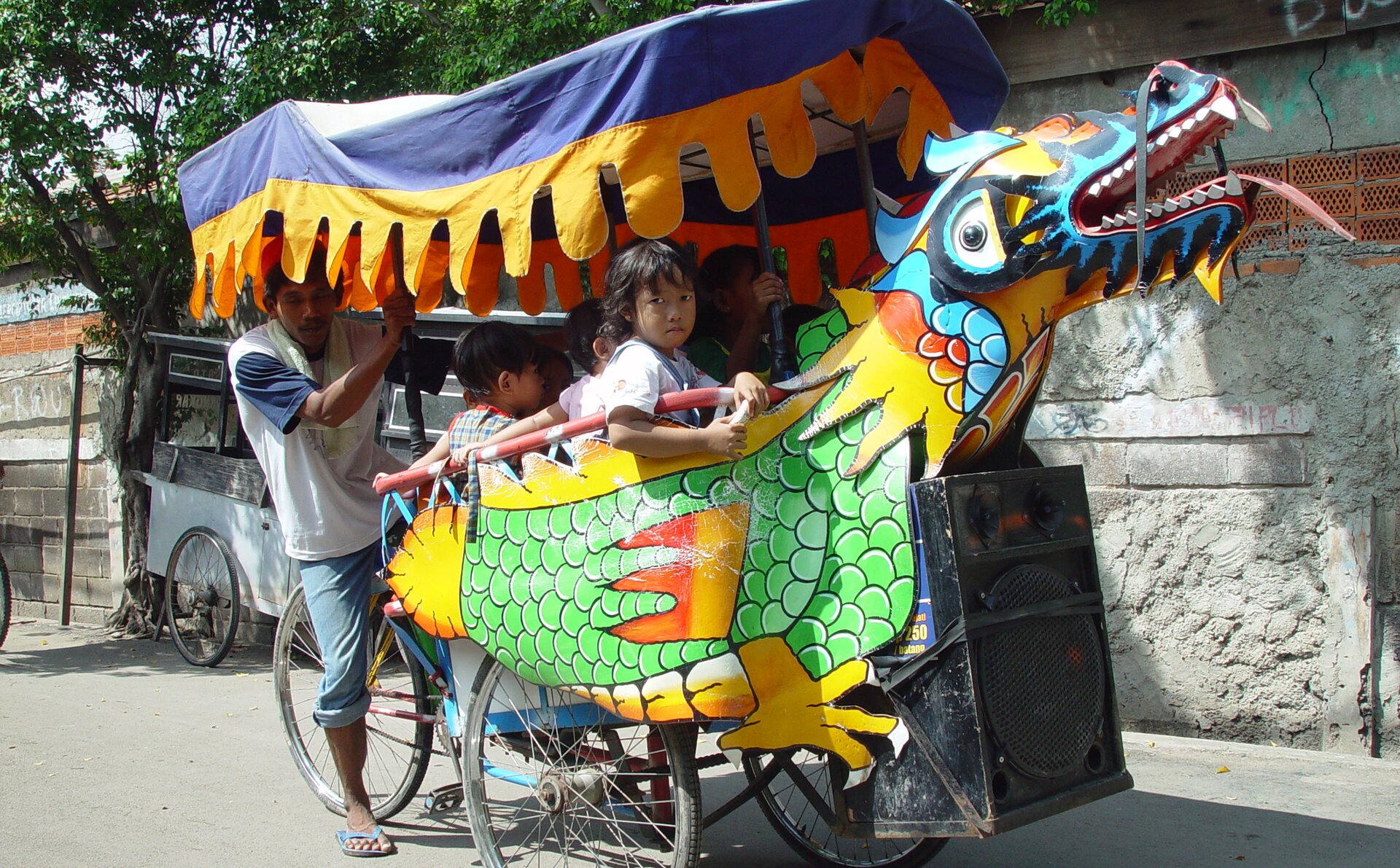 Kliknij, aby powiększyć. Zdjęcie przedstawia indonezyjską rikszę, której kosz ma kształt smoka. Głowa smoka z otwartą paszczą wystaje przed rikszą, a ciało smoka otacza boki rikszy. Smok jest są bardzo kolorowy – zielony, niebieski, żółty. Z paszczy smoka i z jego nozdrzy zwisają kolorowe wstążki. Smok nie wygląda groźnie. Nad rikszą zamocowany jest niebiesko‑żółty baldachim o powycinanych brzegach. W rikszy siedzą dzieci, z tyłu rikszy na siodełku siedzi rikszarz. W tle widać szary mur i stojącą pod nim zwykłą rikszę oraz drzewa.