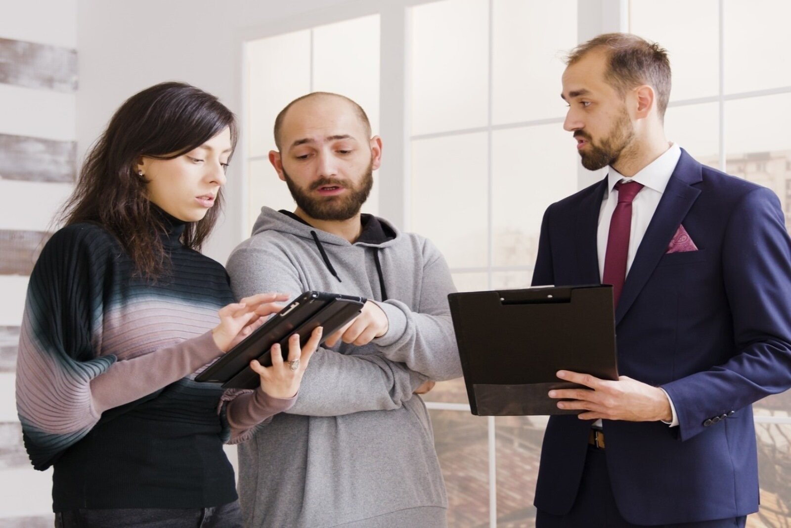 Zdjęcie przedstawia trzy stojące osoby. Po lewej stronie stronie stoi kobieta trzymająca tablet, obok niej mężczyzna w bluzie z kapturem. Oboje spoglądają na ekran tabletu. Po prawej stronie stoi mężczyzna w garniturze, który w dłoniach trzyma teczkę i jest zwrócony twarzą w kierunku kobiety i mężczyzny.