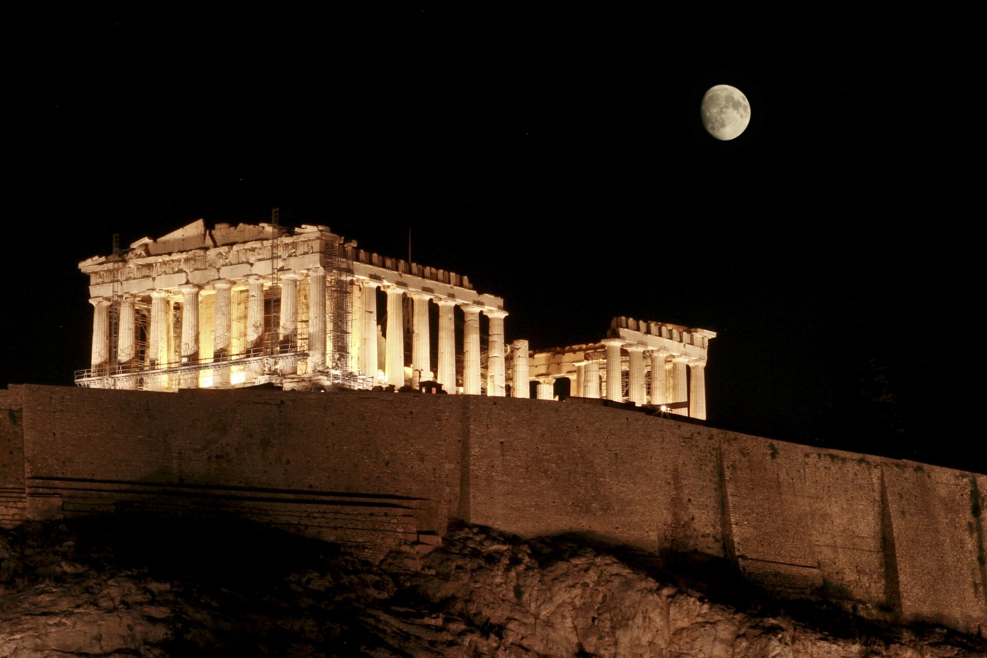 Zdjęcie przedstawia Akropol, na którym widoczny jest Partenon podświetlony światłami w nocy. Zdjęcie zostało zrobione z niższego poziomu, widoczny jest Partenon na wzniesieniu. Niebo jest czarne. Po prawej stronie nad Partenonem widoczny jest prawie cały Księżyc. 