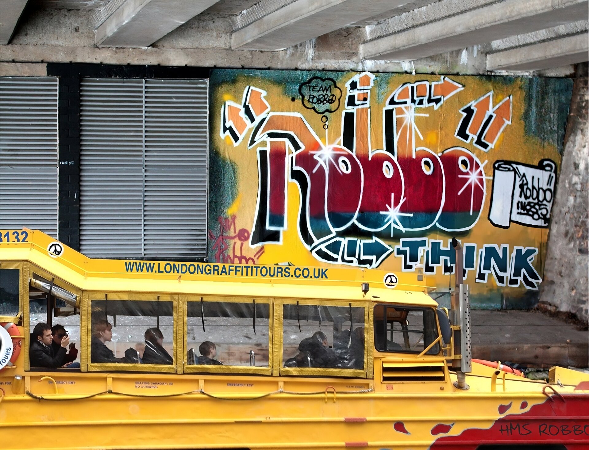 Zdjęcie przedstawia ścianę, znajdującą się pod betonowym zadaszeniem. Na ścianie widoczne są dwa otwory, które przysłonięte są drewnianymi, szarymi, wąskimi belkami. Wokół otworów ściana jest czarna. Obok znajduje się kolorowy napis: "ROBBO". Jest on namalowany na żółto‑miętowym tle. Napis przechodzi z koloru żółtego w czerwony, a z czerwonego w miętowy. Wokół napisu znajdują się strzałki wskazujące w różne kierunki. Obok napisu znajduje się biały pergamin, na którym pojawia się czarny napis"ROBBO". Przed muralem znajduje się żółta łódka, za oknami której siedzą ludzie wpatrujący się wprost przed siebie. Na dachu łódki znajduje się napis "www.londongraffititours.co.uk". Zdjęcie zostało wykonane w świetle dziennym.