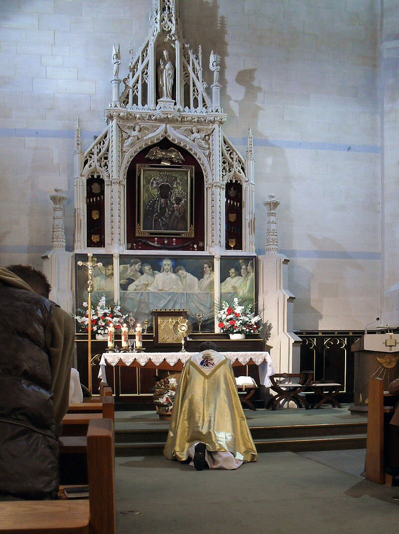 Fotografia przedstawiająca ołtarz kościoła. Jest misternie rzeźbiony w jego centrum znajduje się obraz ukazujący Matkę Boską z Dzieciątkiem. Poniżej obraz przedstawiający Ostatnią Wieczerzę. Przed ołtarzem klęczy duchowny ubrany w złoty ornat.