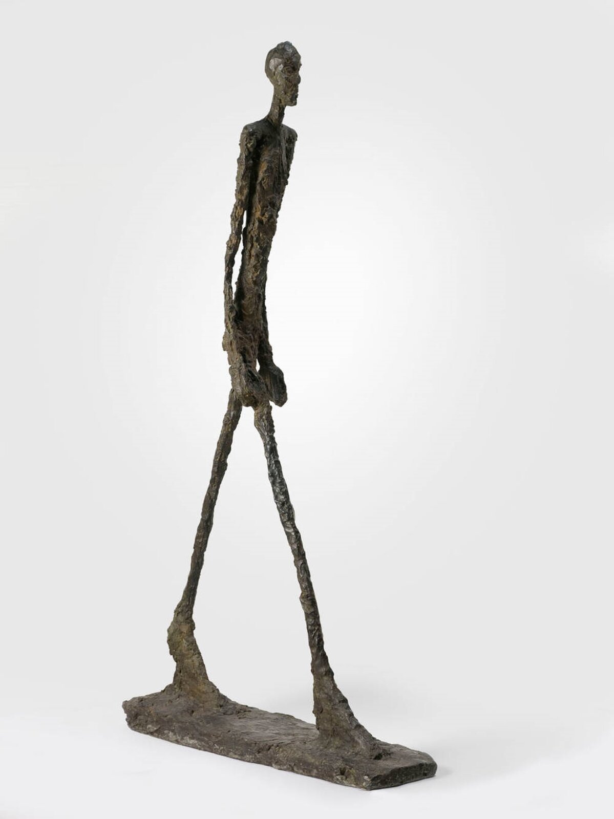 Fotografia przedstawia rzeźbę wykonaną z materiału przypominającego glinę, uchwyconą na białym tle. Obecna jest prostokątna podstawa oraz umieszczona na niej figura o nieproporcjonalnie długich i szczupłych nogach oraz rękach, a także dużych stopach. Nogi postaci są w rozkroku, jak gdyby się poruszała. Jej ręce są opuszczone wzdłuż ciała, którego tors jest lekko pochylony. Plecy są proste, a twarz skierowana do przodu.