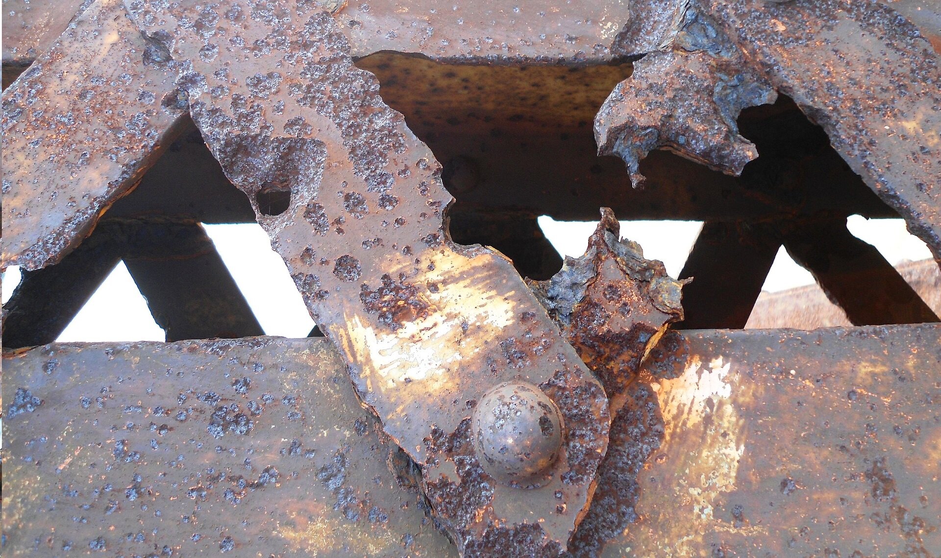 Zdjęcie przedstawia fragment metalowej konstrukcji mostu. Przedstawione elementy są silnie skorodowane. Na ilustracji są przedstawione liczne ubytki w warstwie metalu, wżery oraz perforacje.