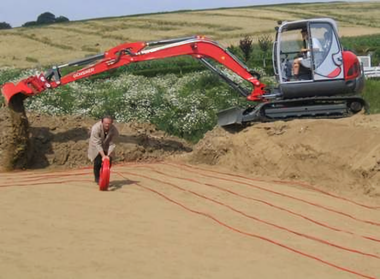 Na zdjęciu przedstawiono układanie rur kolektora do pomp z bezpośrednim odparowaniem. Na pierwszym planie widać mężczyznę rozprowadzającego przewody na odkrytej i wyrównanej powierzchni. W tle widoczna jest koparka.