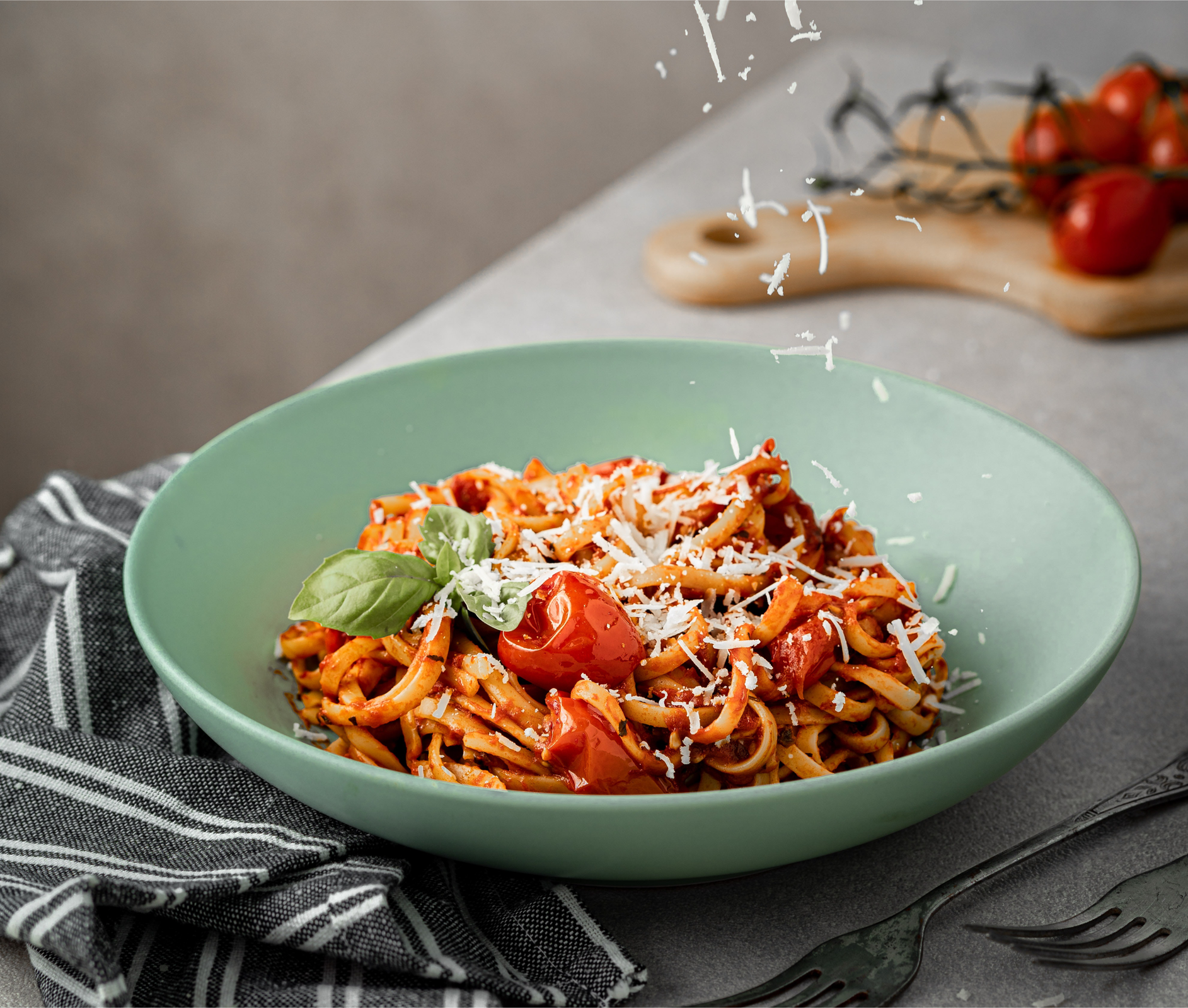 Zdjęcie przedstawia włoskie danie, makaron z pomidorami. Danie jest posypane serem i przybrane listkami bazylii. Talerz stoi na blacie. W tle widoczna jest drewniana deska kuchenna, na której leży gałązka pomidorów koktajlowych. Obok talerza leży ściereczka kuchenna w paski oraz dwa widelce.