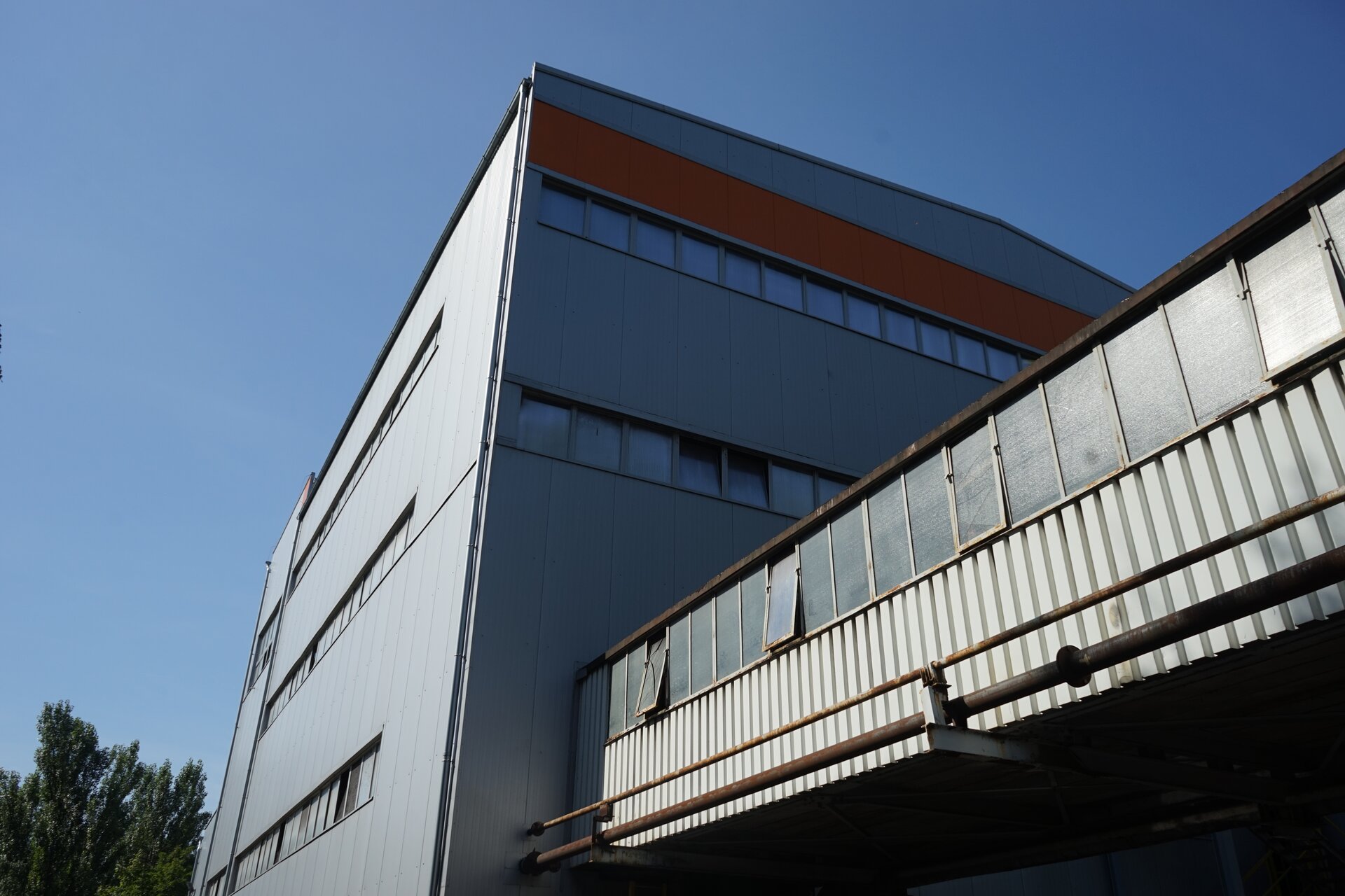 Na zdjęciu widoczny jest most transportowy. Po lewej stronie zdjęcia widoczny jest duży prostopadłościenny budynek. Budynek jest w kolorze szarym. Do podstawy budynku , poniżej pierwszego rzędu okien, przyłączony jest łącznik. Łącznik wykonany jest z szarej blachy falistej. W górnej części ściany osadzone są okna. Jest to most transportowy.Na terenie placu widać części urządzeń oraz metalową konstrukcję przypominającą ramę przenośnika.