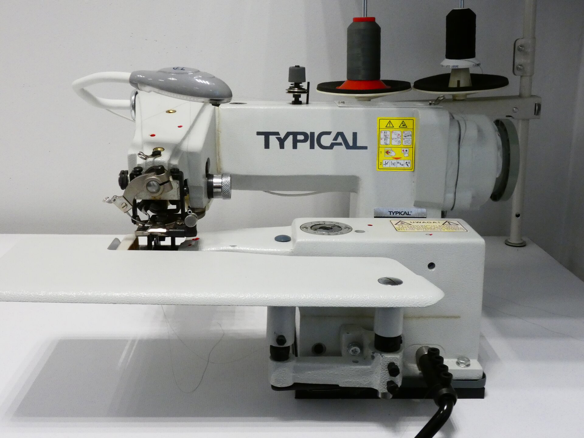 Zdjęcie przedstawia białą maszynę z napisem TYPICAL. Maszyna składa się z ramienia z igłą, z tyłu zaś są widoczne dwie ciemne nici nawinięte na szpule. 