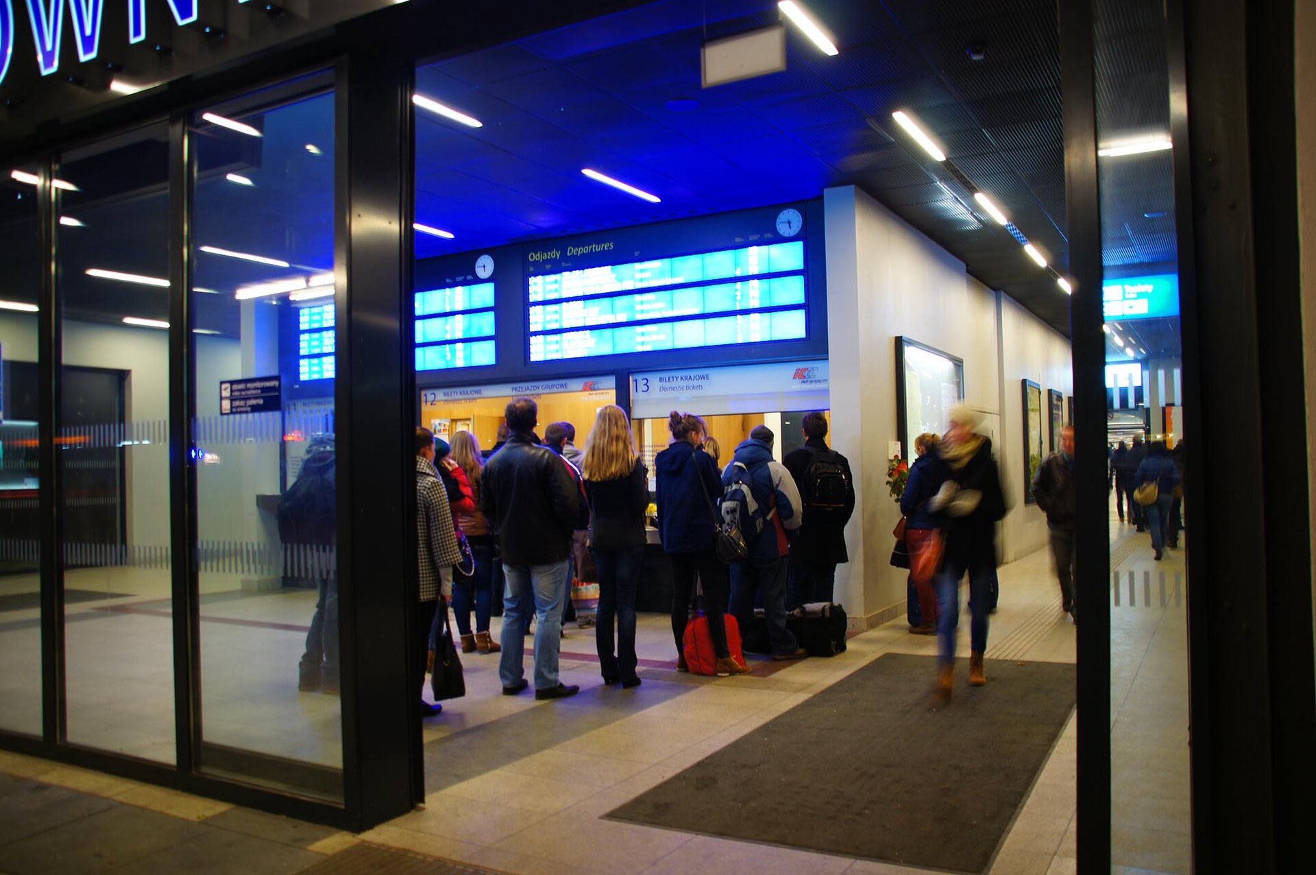 Fotografia przedstawiająca dwie kasy biletowe: dwunastą i trzynastą na Dworcu Głównym we Wrocławiu. Przy kasach stoją kolejki. Ludzie są zwróceni frontem do kas. W kolejce do kasy numer trzynaście stoi sześć osób. Kolejka do kasy dwunastej jest częściowo zasłonięta. Na prawo od kas widać fragment holu dworca, przez który idą ludzie.
