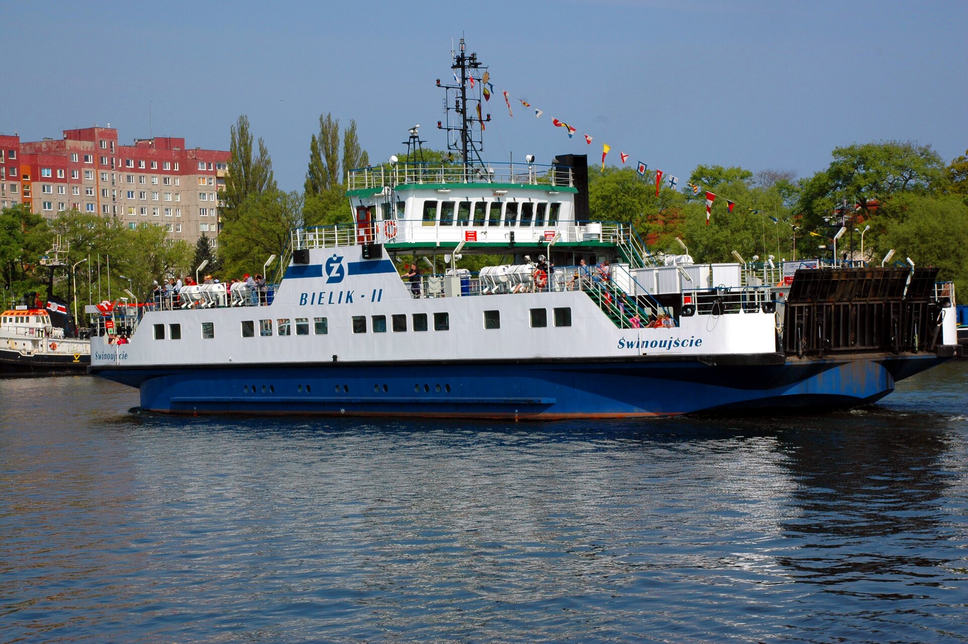 Zdjęcie promu. Ma długość około dwudziestu metrów, wzdłuż burty rząd okien, pasażerowie na pokładzie. W tle linia drzew i zabudowania miejskie.