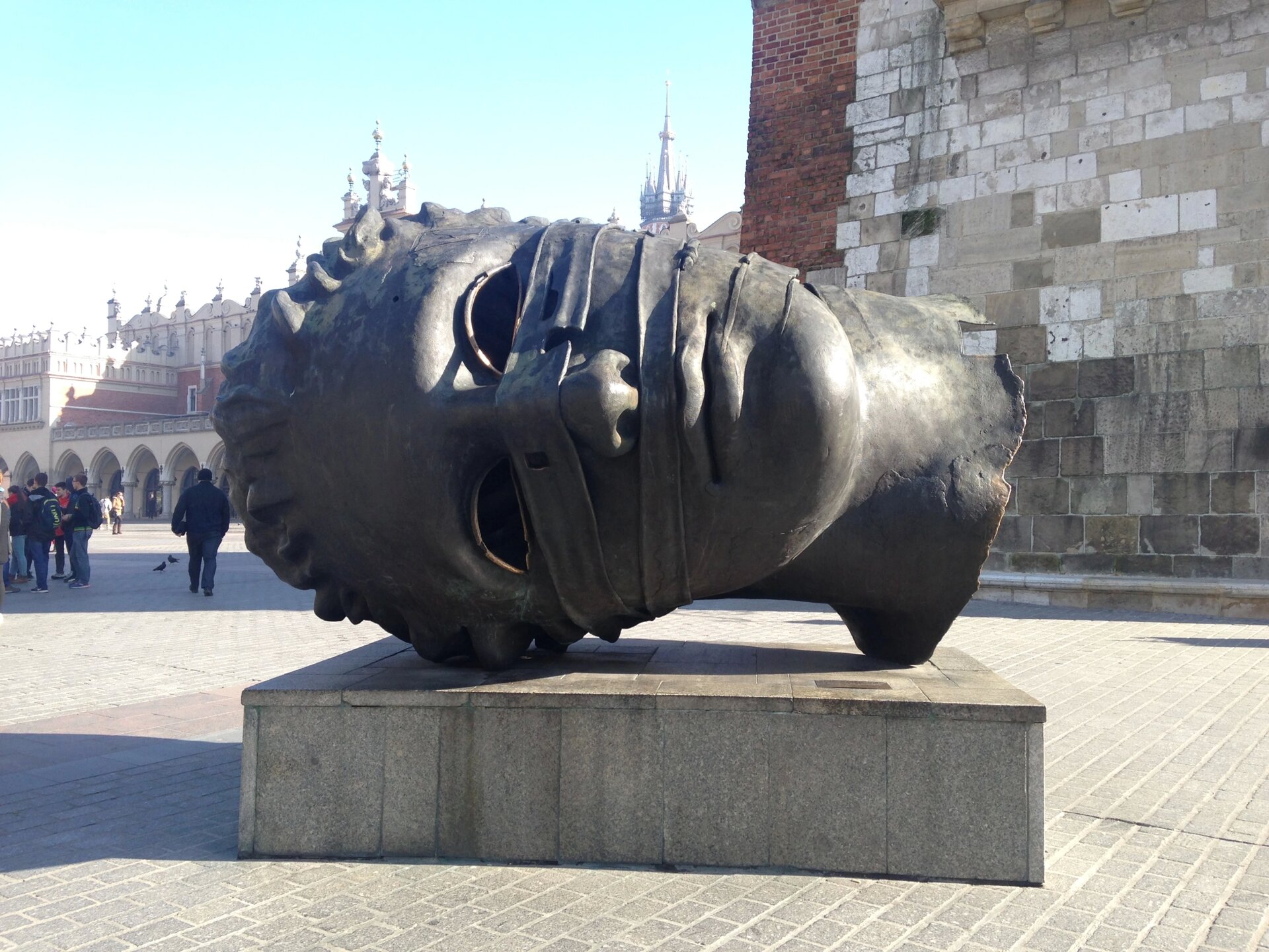 Zdjęcie przedstawia rzeźbę w kolorze szarym. Przedstawia dużą, leżącą głowę. Z lewej strony są włosy, a z prawej szyja. Rzeźba ma kręcone włosy, otwory w miejscach gałek ocznych i zamknięte usta. Przez grzbiet nosa i pod nosem przechodzą 2 paski. Szyja ma nierówną fakturę i krawędzie. Rzeźba jest na podeście wykonanym z szarych, prostokątnych płyt. Dookoła jest szara kostka. W tle, z prawej strony jest ściana z białych, szarych i czerwonych cegieł o różnej wielkości. W tle, z lewej strony są ludzie w ciemnych ubraniach i fragment beżowo‑czerwonego budynku z wieżyczkami. 