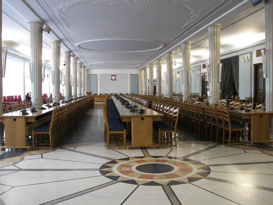 Zdjęcie przedstawia dużą salę z dwoma szeregami kolumn, w której znajdują się trzy rzędy stołów z krzesłami. Na końcu sali wisi na ścianie polskie godło.