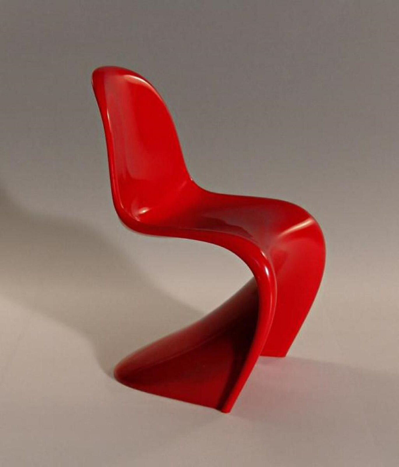 Fotografia przedstawia krzesło Panton projektu Vernera Pantona – krzesło jest czerwone, wykonane z jednego kawałku plastiku, odlewane w formie, nóżki krzesła są płytkie, cofnięte do tyłu. Zdjęcie zostało wykonany na szarym tle. 