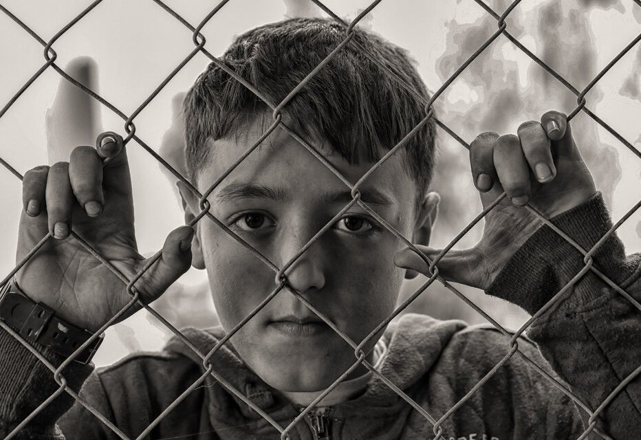 Zdjęcie czarno-białe przedstawia chłopca trzymającego się płotu z siatki metalowej, ma on krótkie włosy, a ubrany jest w bluzę z kapturem. Patrzy w obiektyw z powagą, w tle rozmazane drzewa.