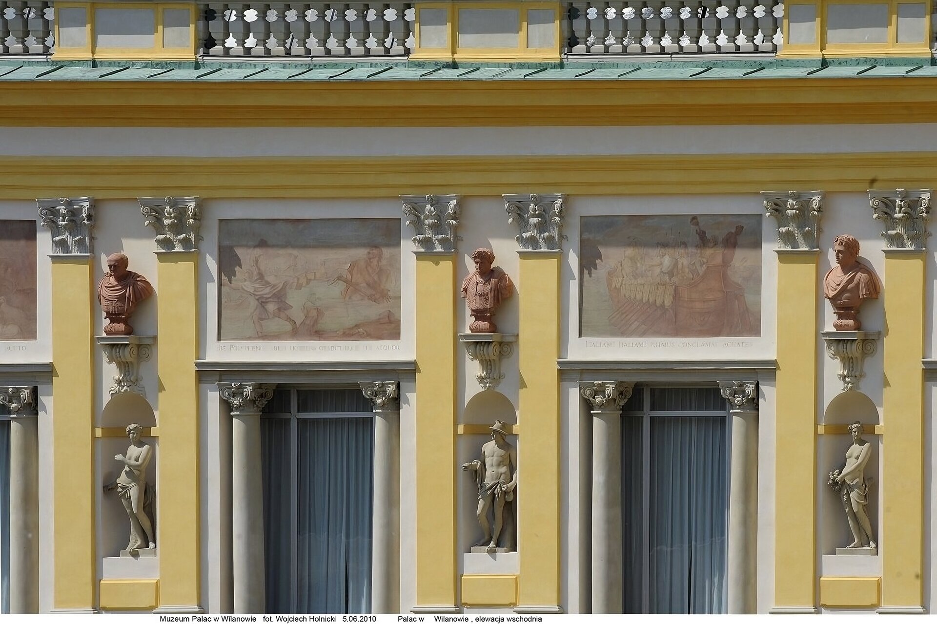 Zdjęcie przedstawia jasnożółtą elewację budynku.  Dwa duże prostokątne okna z białymi firankami i kolumnami po bokach. Pomiędzy oknami znajdują się ozdobne rzeźby. Nad oknami umieszczone są kolorowe freski. 