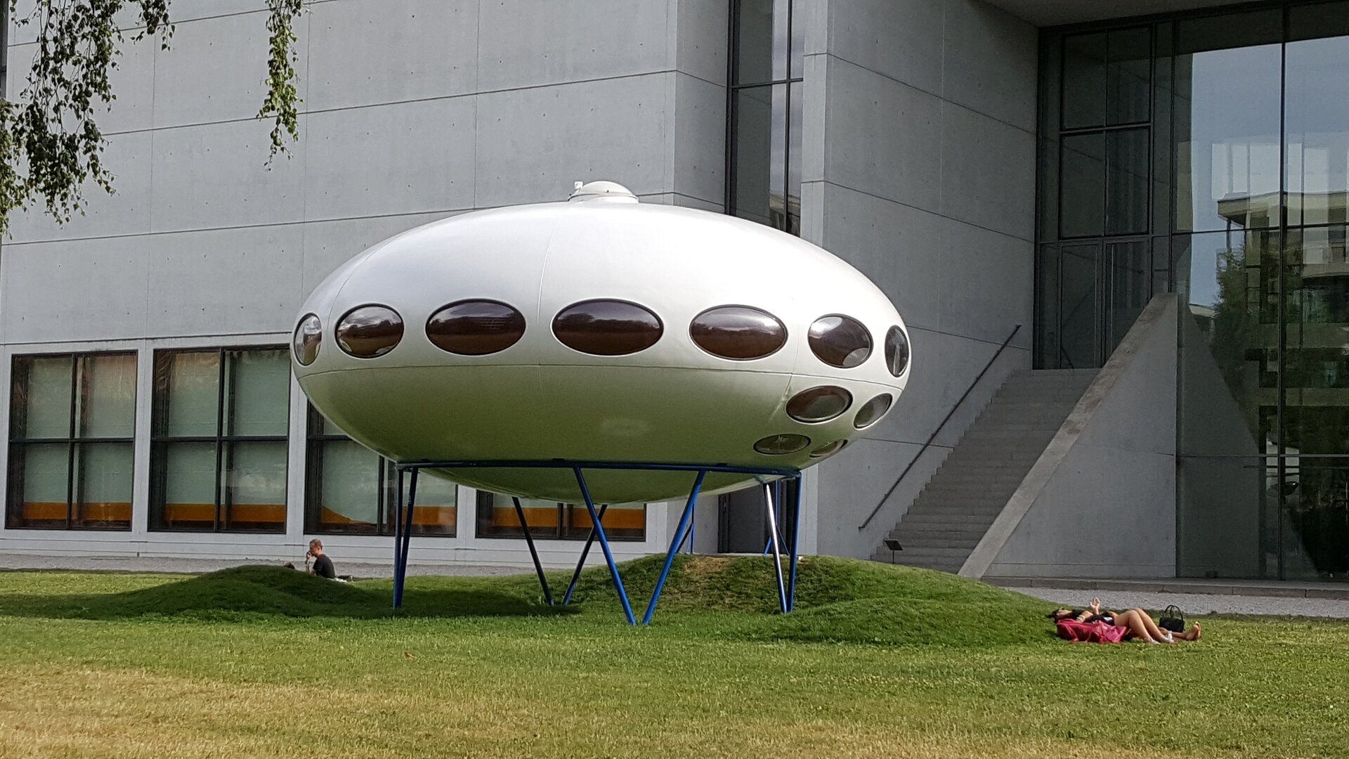 Ilustracja przedstawiająca dom futuro w Monachium. Ma on kształt elipsoidy z oknami wzdłuż obwodu w kształcie eliptycznym. Budynek ustawiony jest na metalowym stelażu. W tle znajdują się inne zabudowanie w współczesnym stylu. Obok na trawie leżą ludzie.