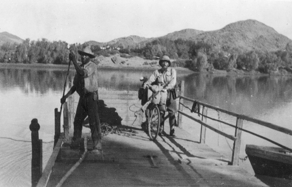 Zdjęcie przedstawia przeprawę niewielkim, drewnianym promem przez rzekę. Na promie znajduje się czarnoskóry wioślarz oraz biały mężczyzna z rowerem.