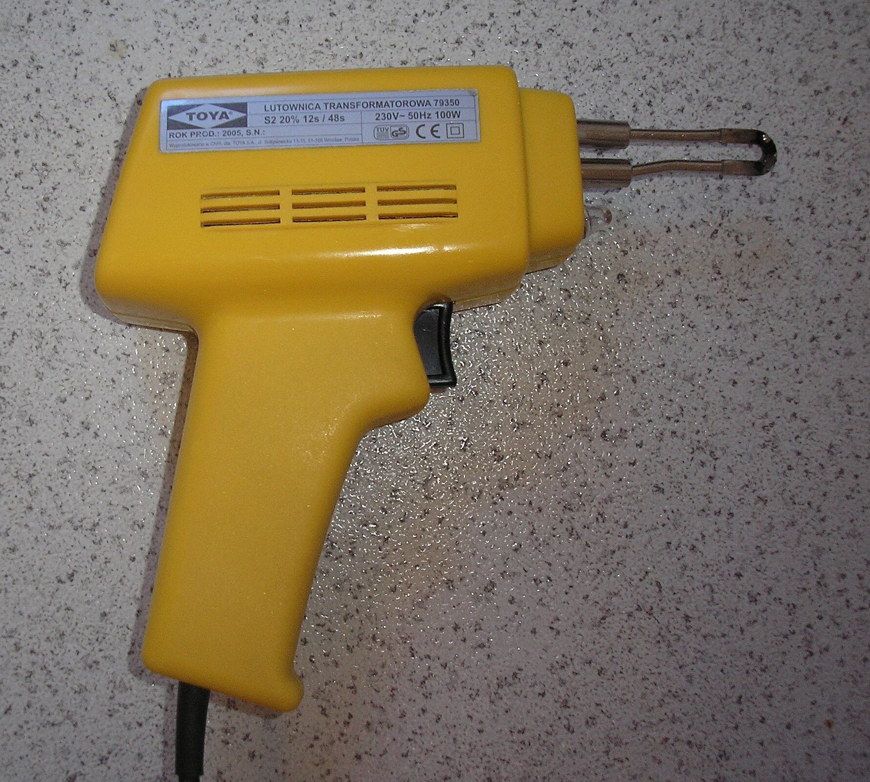 Zdjęcie przedstawia lutownice transformatorową, wyglądającą jak plastikowy pistolet, z którego wystaje kabel na dole rękojeści. W górnej części poziomo ustawiona jest grzałka.