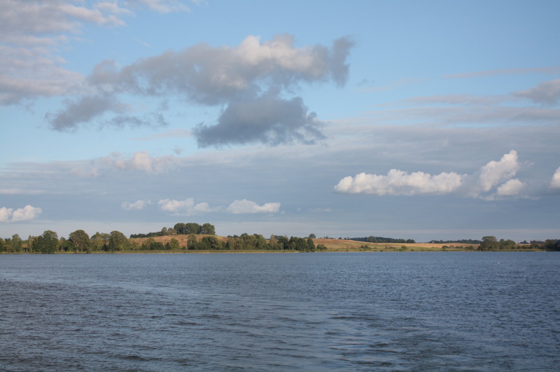 Zdjęcie przedstawia jezioro Niegocin. Na pierwszym planie widać lustro wody, dalej przeciwny brzeg. Na nim znajdują się wzgórza i drzewa.