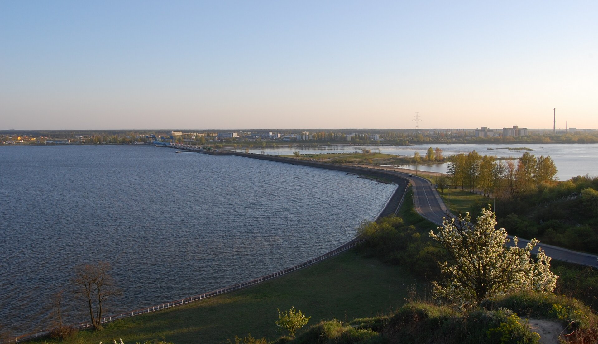 Fotografia przedstawia zaporę wodną, na niej szosa. Po lewej stronie zapory spiętrzona woda, po prawej rozlewiska. W oddali miasto.