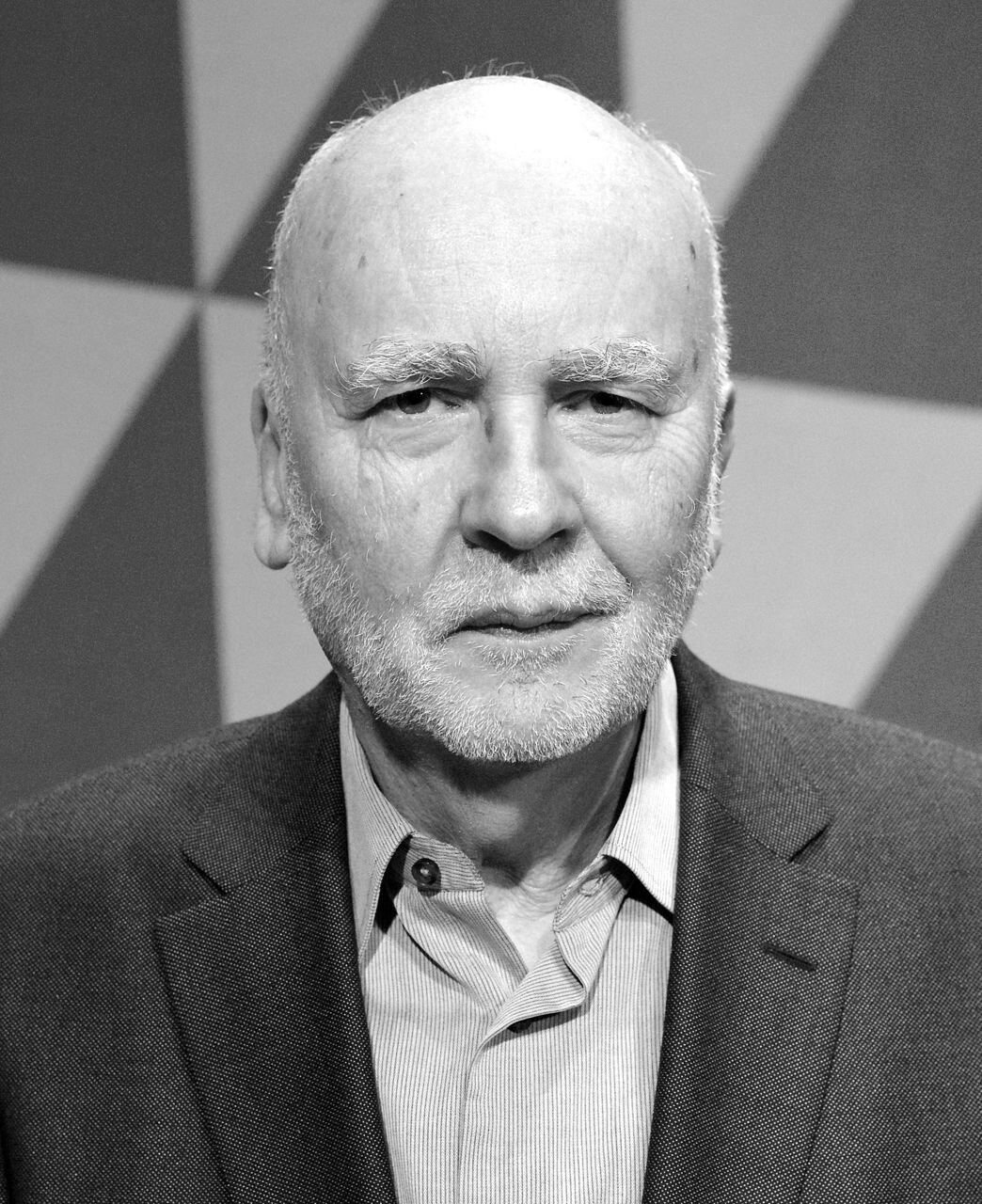 Zdjęcie przedstawia portret starszego mężczyzny. Ma podłużną twarz ze zmarszczkami, jest łysy i posiada krótką, siwą brodę z wąsami. Lekko się uśmiecha. Ubrany jest w ciemną marynarkę z jaśniejszą koszulą pod spodem. Tło zdjęcia składa się z ostrych trójkątów.