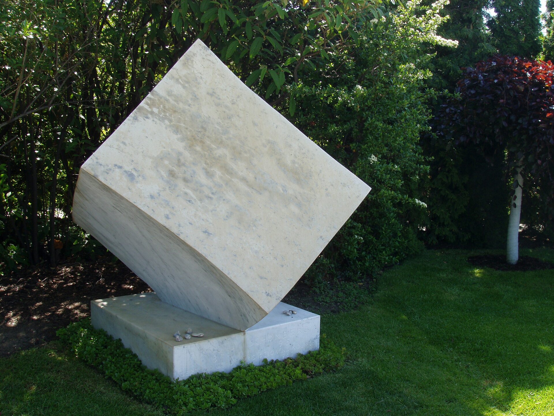 Fotografia przedstawia grób Schönberga w Wiedniu. Nagrobek z marmuru w kształcie sześcianu ustawionego na jednym z narożników. Dookoła zieleń.